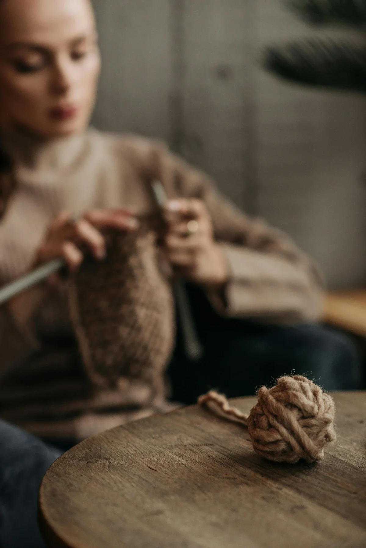Chunky wool yarn ball rests on a table while a woman knits softly blurred in the background of a cozy indoor scene.