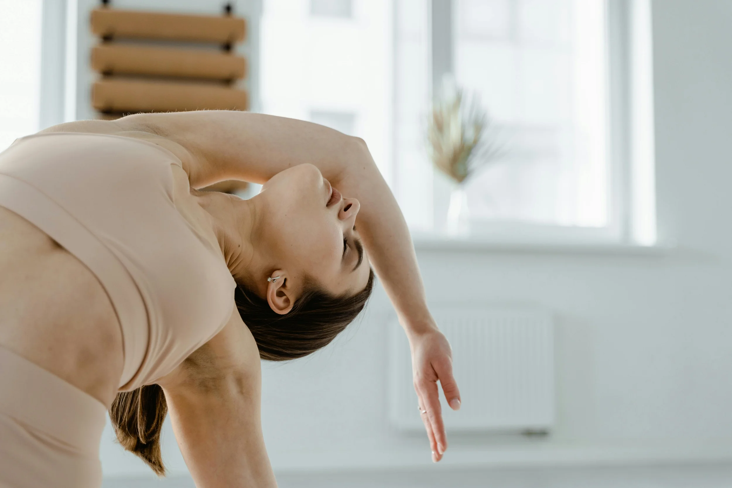 Woman in neutral-tone sportswear performing a deep back-bend stretch in a bright, minimalist Pilates studio.