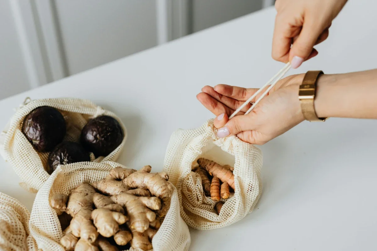 Person cinching a reusable mesh produce bag of turmeric beside bags of ginger and passionfruit on a white countertop.