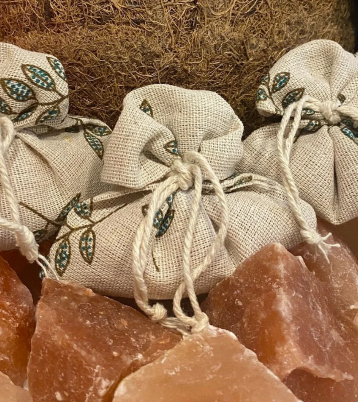  Small embroidered linen drawstring sachets nestled among rough pink Himalayan salt chunks and coconut fibers. 