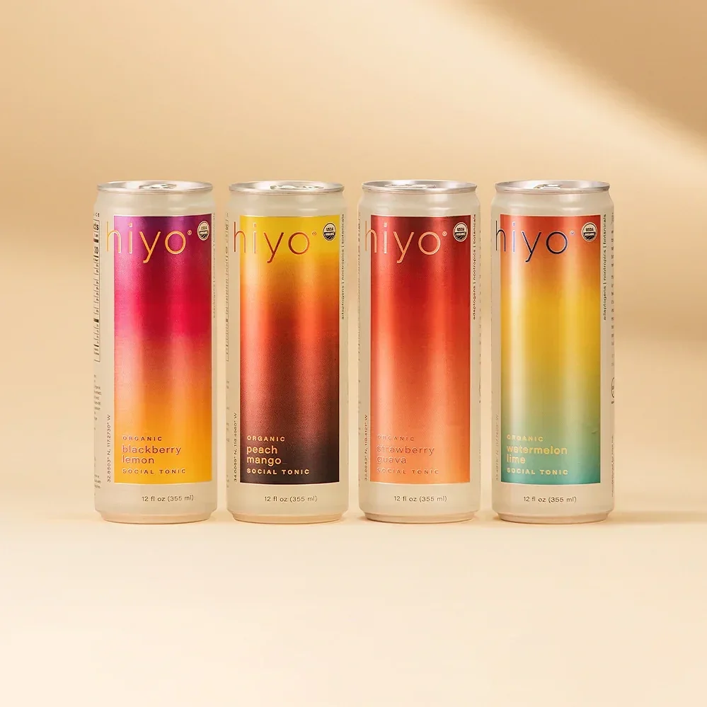  Four tall Hiyo social tonic cans, blackberry lemon, peach mango, strawberry guava, watermelon lime, lined up against a soft beige backdrop. 