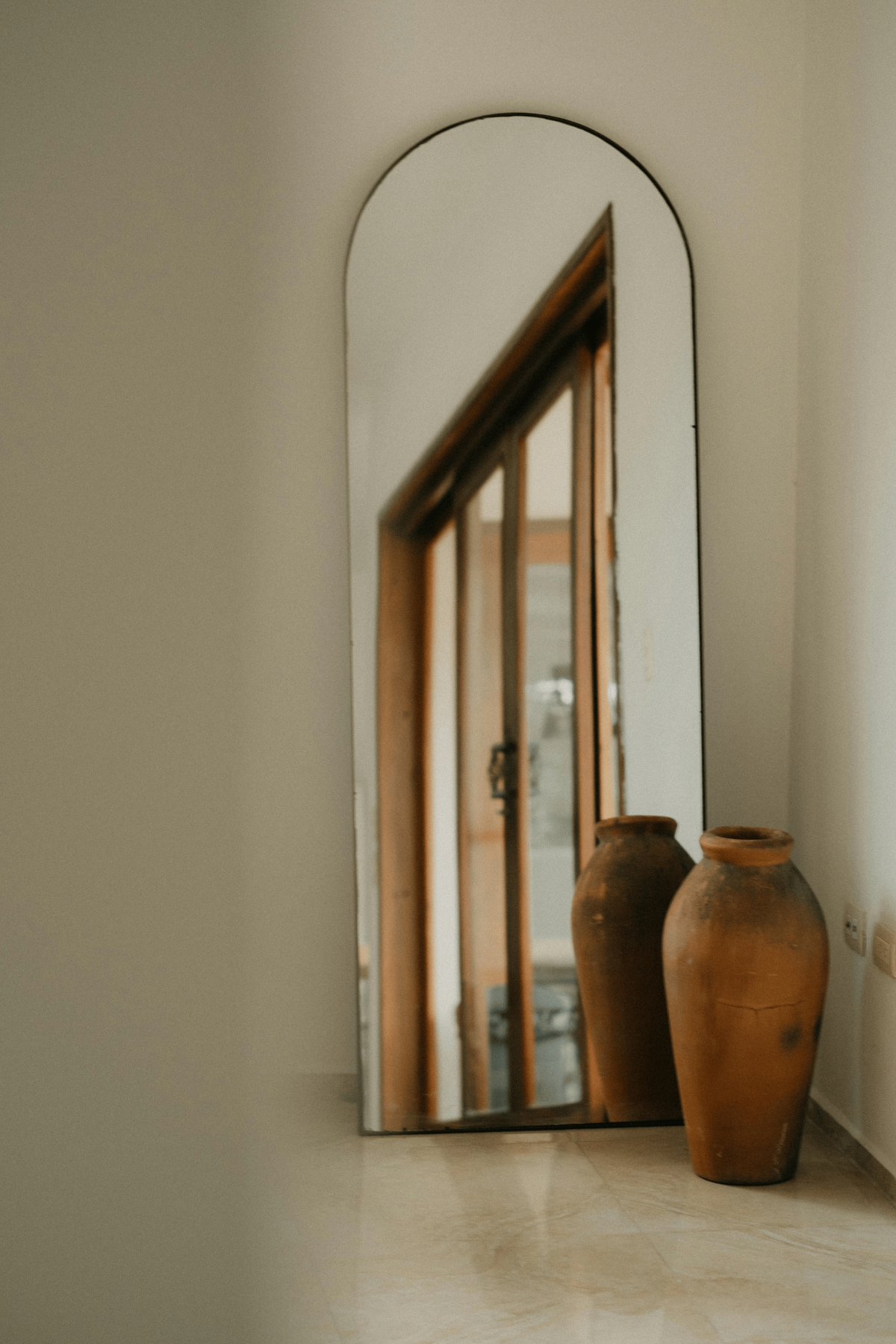 Tall arched mirror reflecting hallway, styled with rustic terracotta urns for Mediterranean interior.