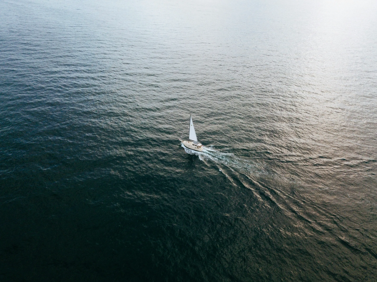 Aerial view of lone white-sail yacht carving gentle wake across open deep-blue ocean (concept image, not Orient Express Corinthian).
