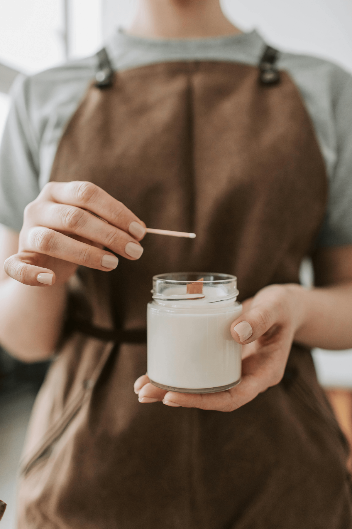 Woman in brown apron prepares to light handcrafted white soy candle with wooden wick, sustainable craftsmanship.