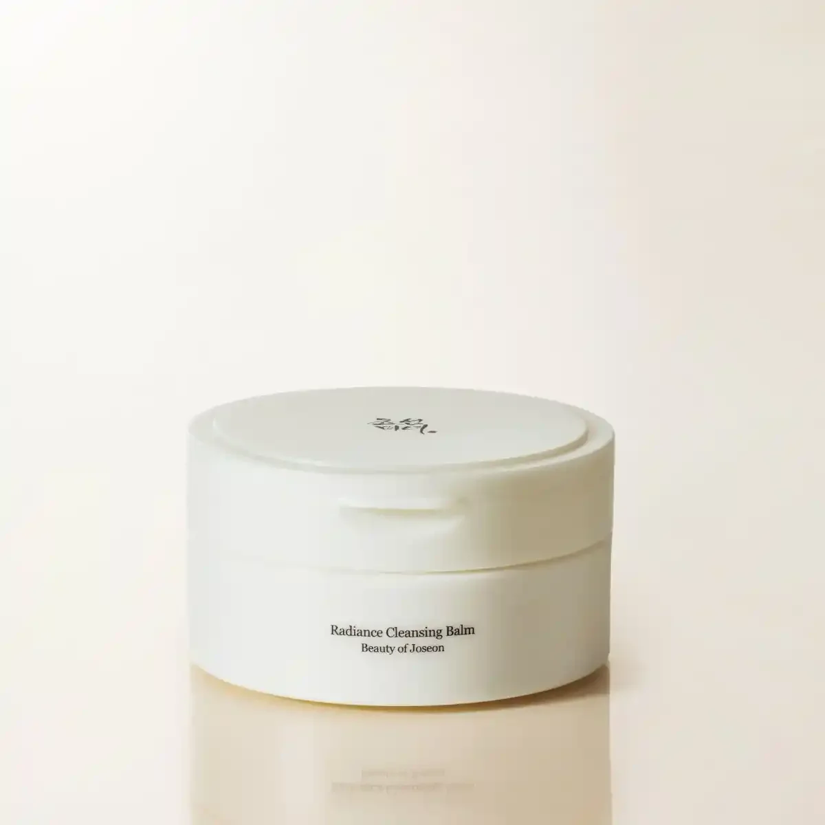 Minimal white tub of Beauty of Joseon Radiance Cleansing Balm shot on a soft beige reflective surface.