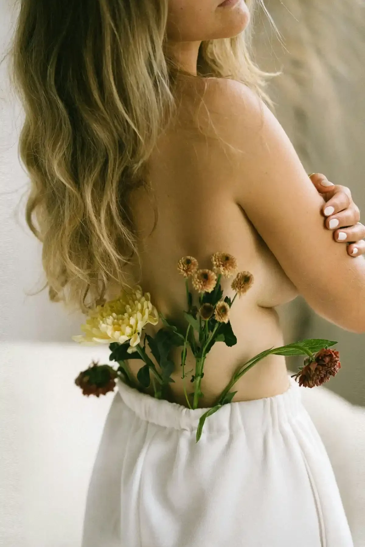 Side view of topless woman with wavy blond hair; fresh flowers tucked into the waistband of white trousers.