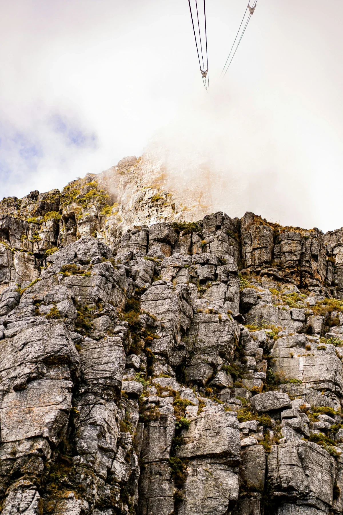 Steep rocky cliffs of Table Mountain with cable car lines disappearing into mist above the summit.