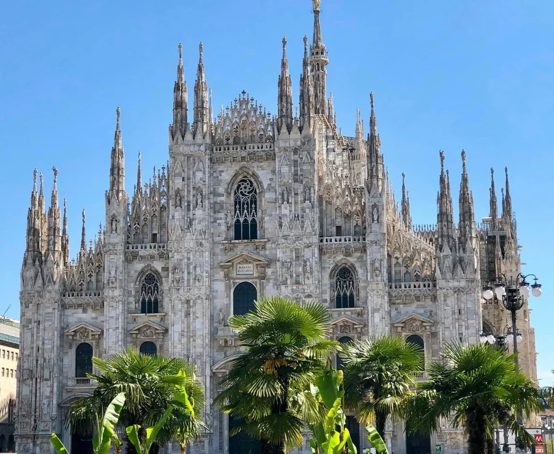  Ornate Gothic marble façade of Duomo di Milano framed by palm trees against a clear blue sky. 