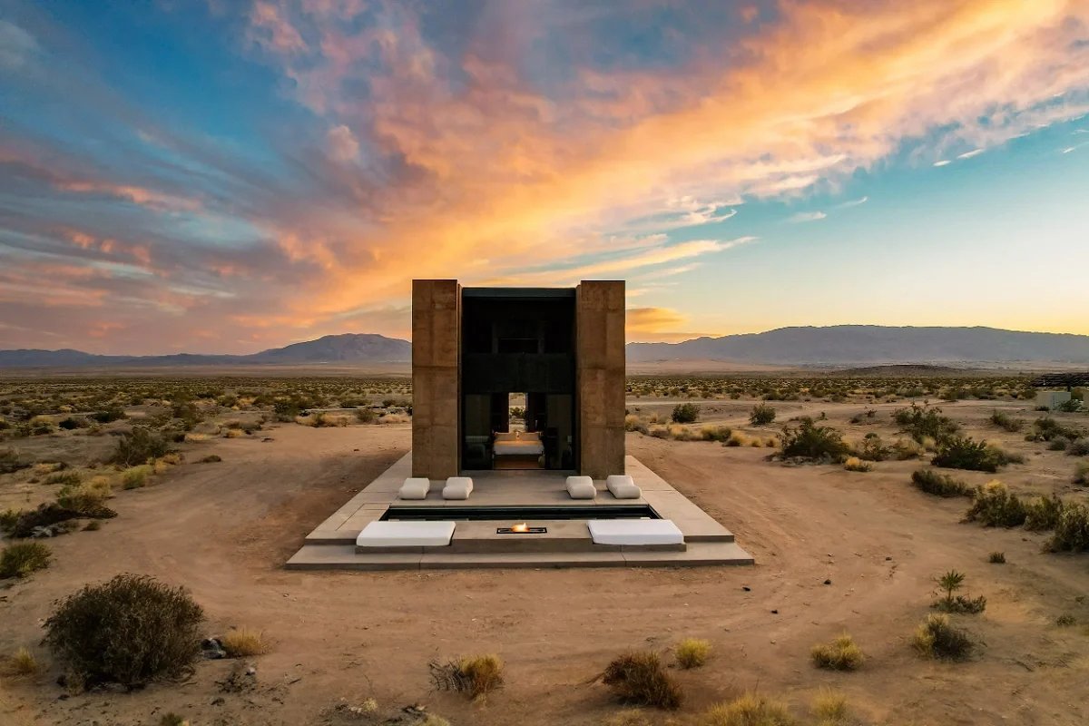  Minimalist concrete cabin glows against a vast Mojave Desert plain beneath a dramatic pink-orange sunset sky. 