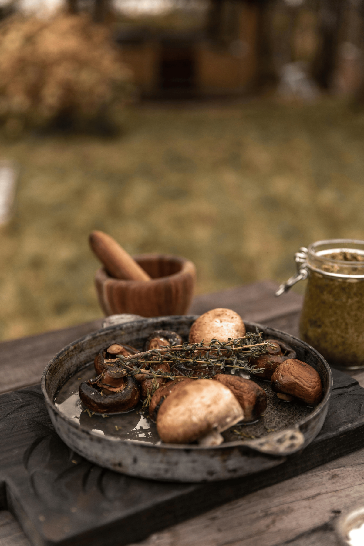 Cast-iron pan of thyme-roasted button mushrooms with pesto jar on wooden picnic table in garden setting.