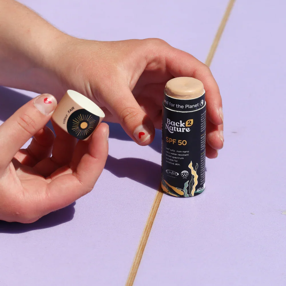 Hands open reef-safe SPF 50 zinc sunscreen stick from Back 2 Nature on lilac surface.