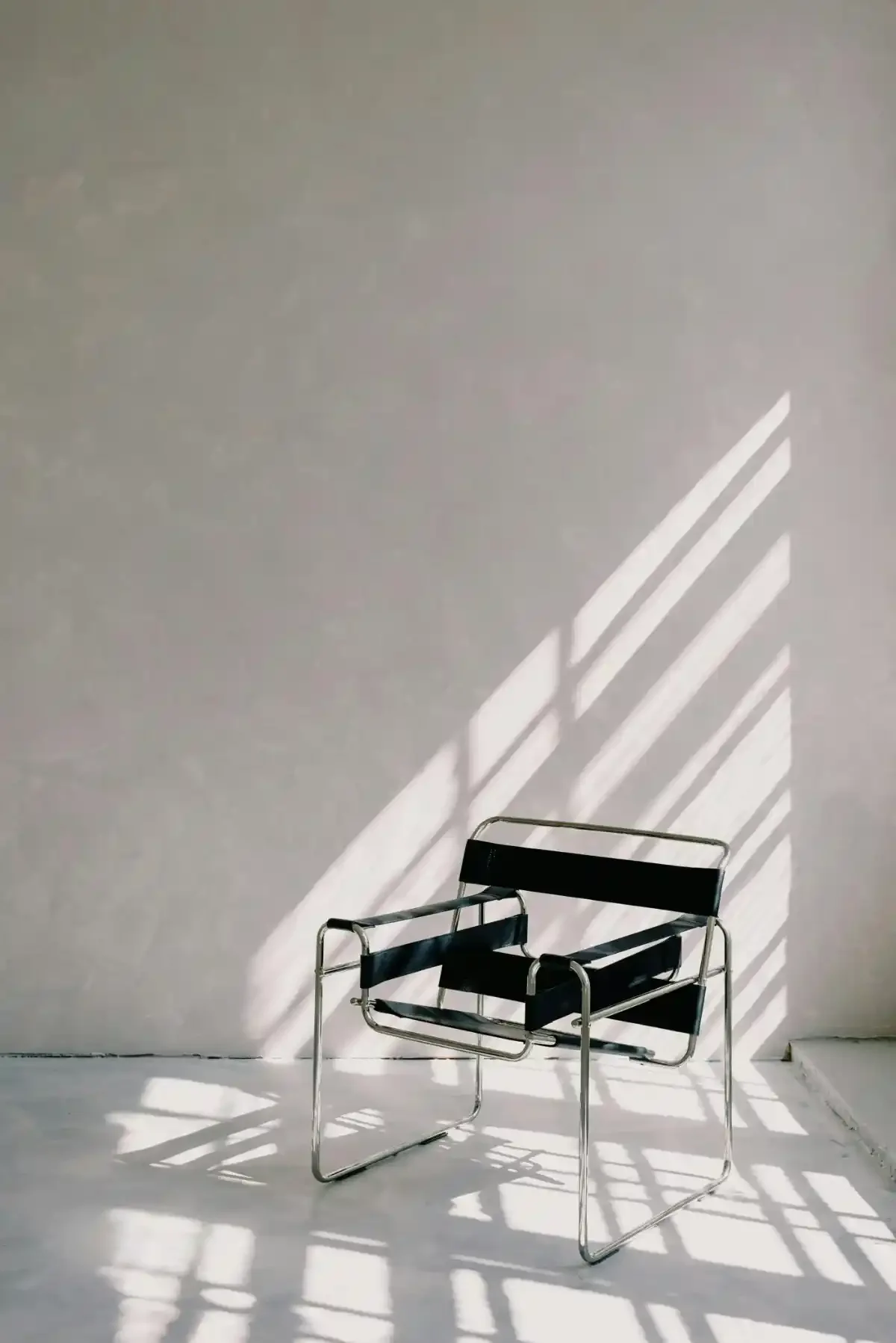 Lone black Wassily chair bathed in diagonal window-light shadows in a sparse white studio.