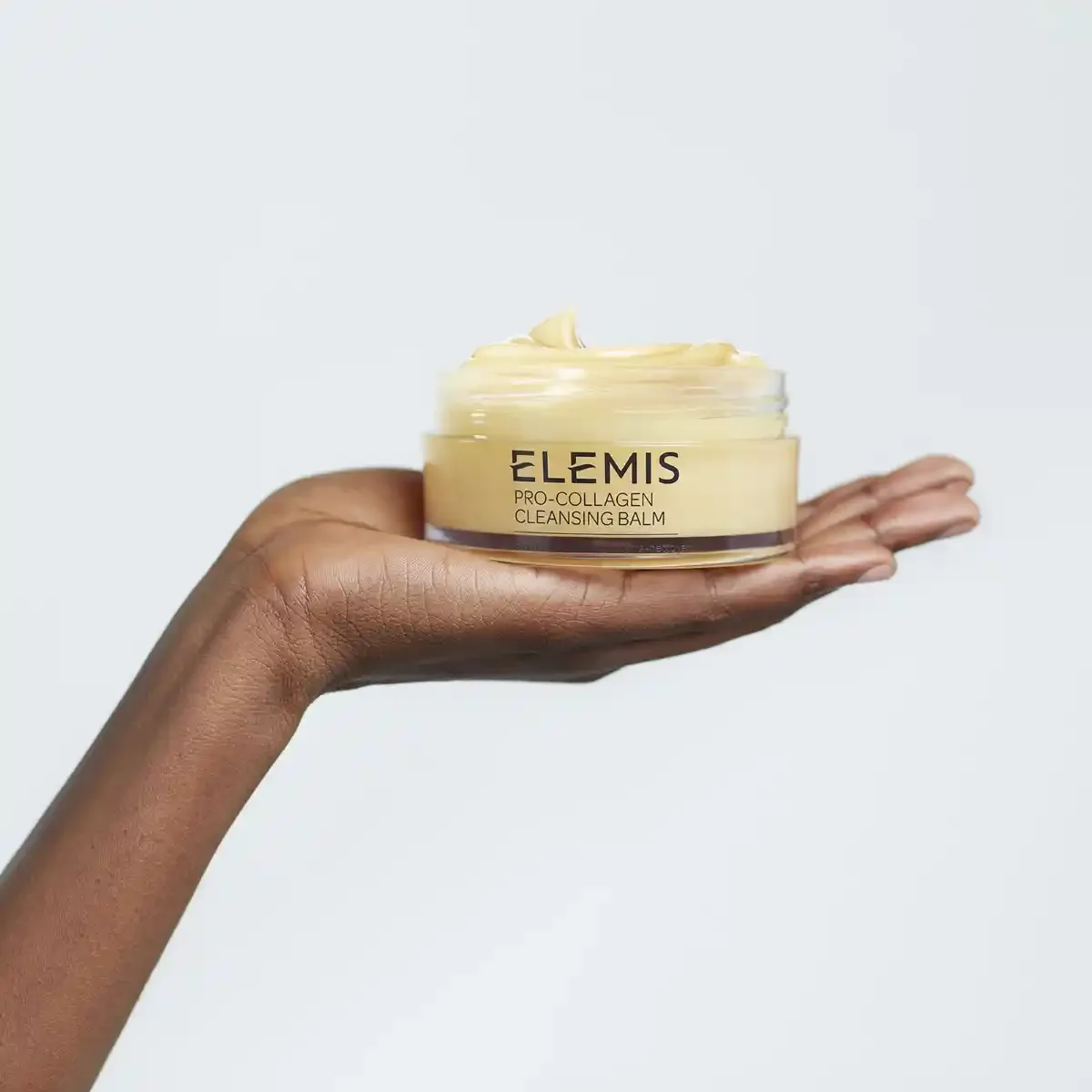 Hand holding a jar of Elemis Pro-Collagen Cleansing Balm against a light grey backdrop.