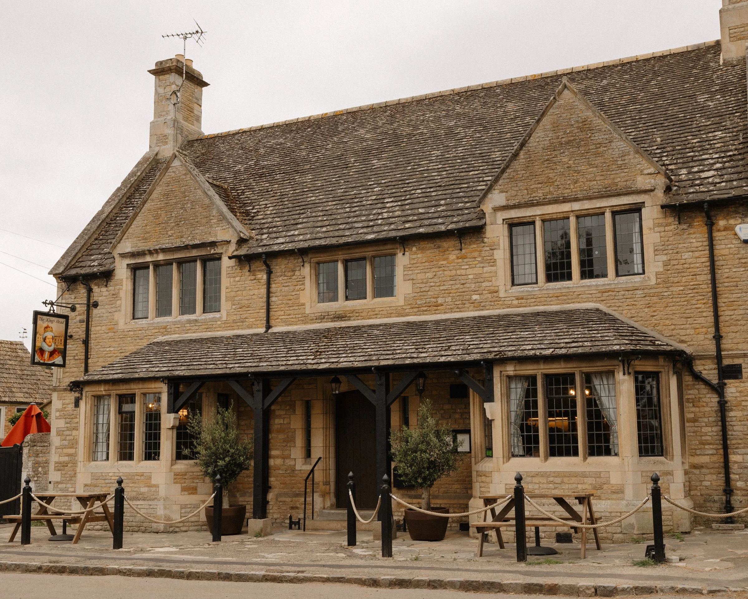 Stone building with multiple windows, a small porch with benches, and a sign with a king's face outside, resembling a historical pub or inn.