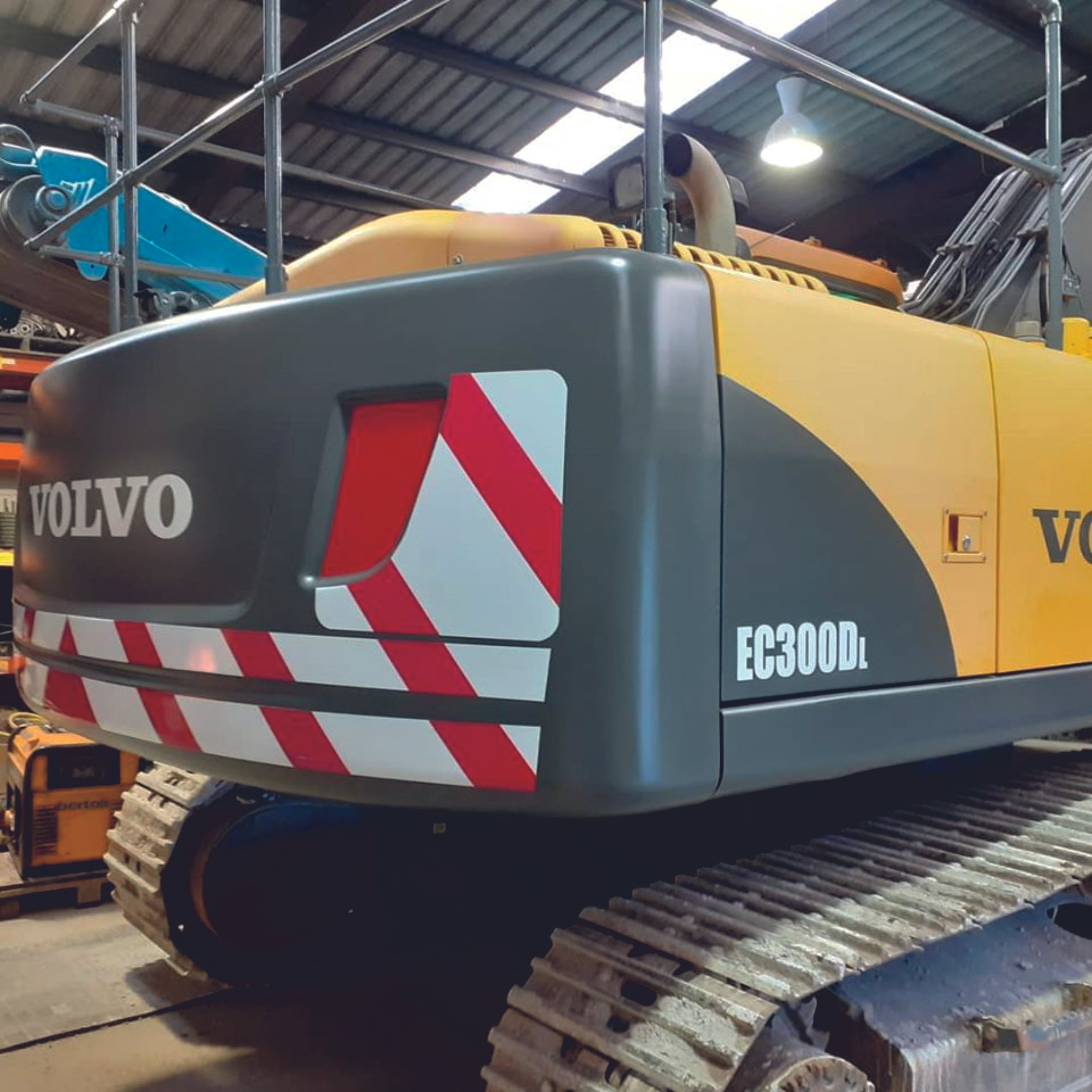Back view of a yellow and black Volvo EC300DL excavator with red and white diagonal stripes, parked indoors. Vehicle wrap. Vehicle Graphics. Commercial Vehicle Branding. Plant Hire Signage. Signmakers Cork & Kerry, Ireland.