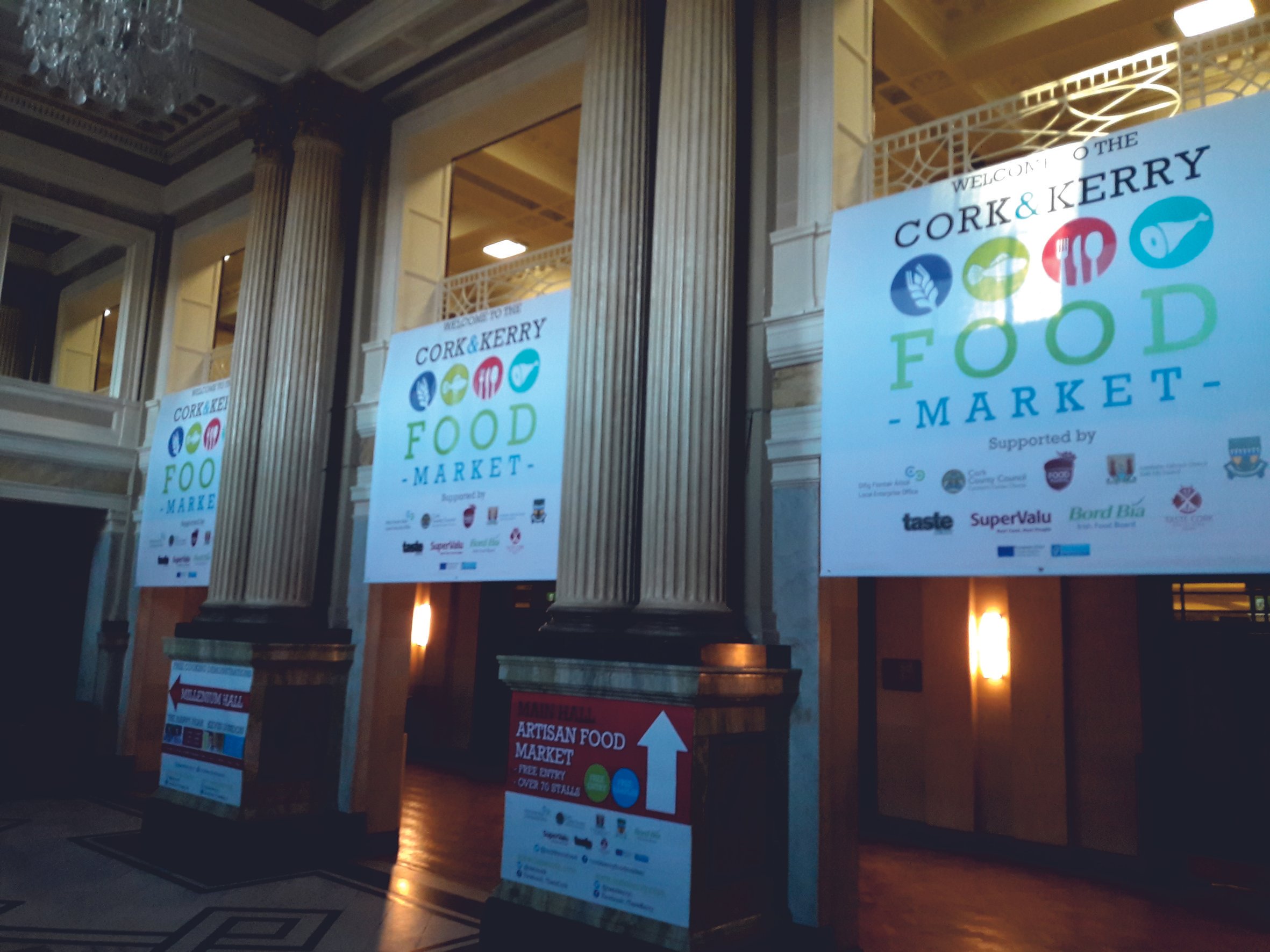 Interior of a building with large posters advertising the Cork & Kerry Food Market, supported by various organizations. The posters are placed on ornate pillars, with visible signage for artisan food markets. Signmakers Cork & Kerry, Ireland.