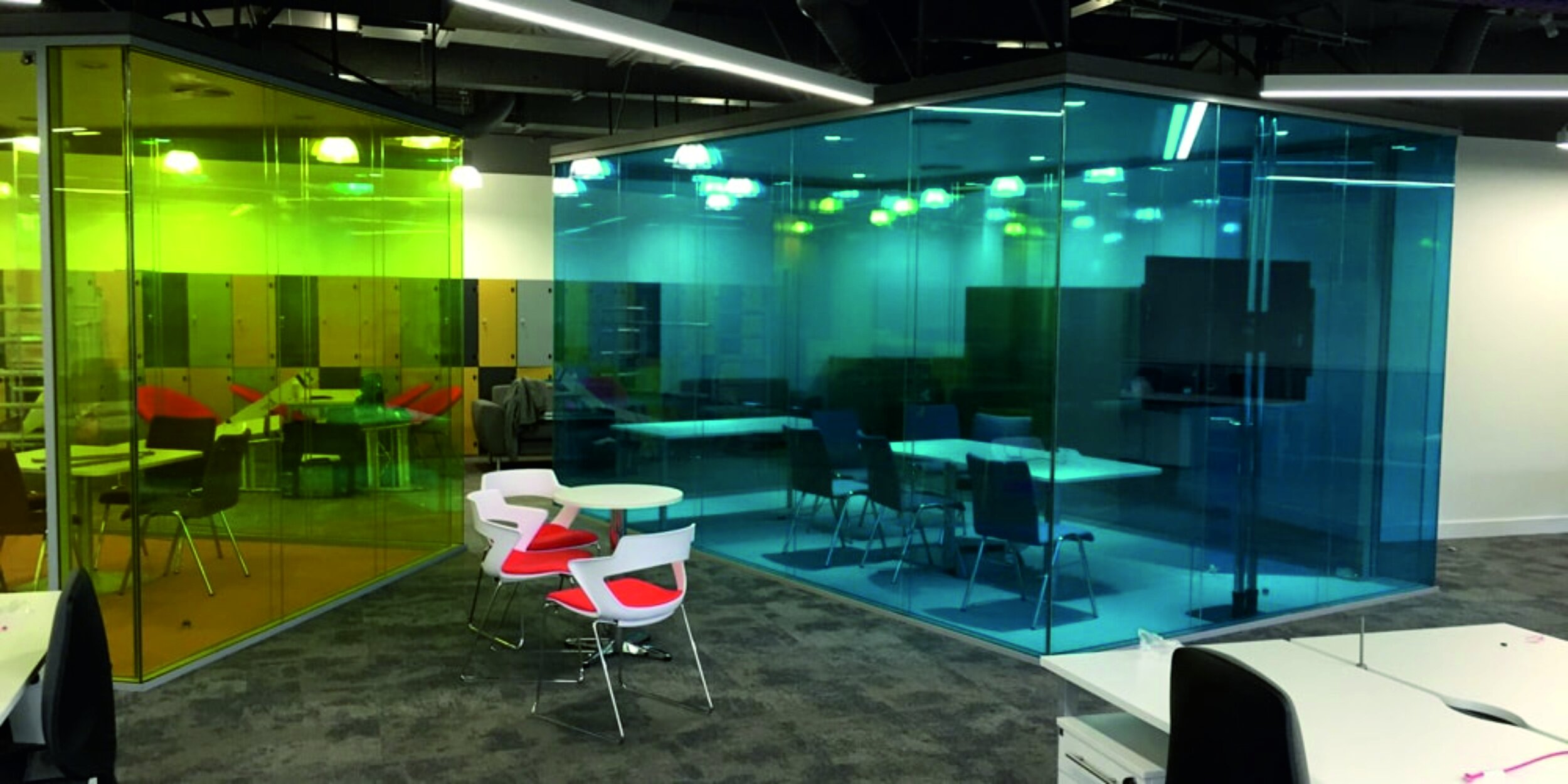 Modern office space with colorful glass meeting rooms, featuring yellow and blue tinted glass walls, and contemporary furniture including white and red chairs around tables. Window manifestations. Printed Film. Signmakers Cork & Kerry, Ireland.