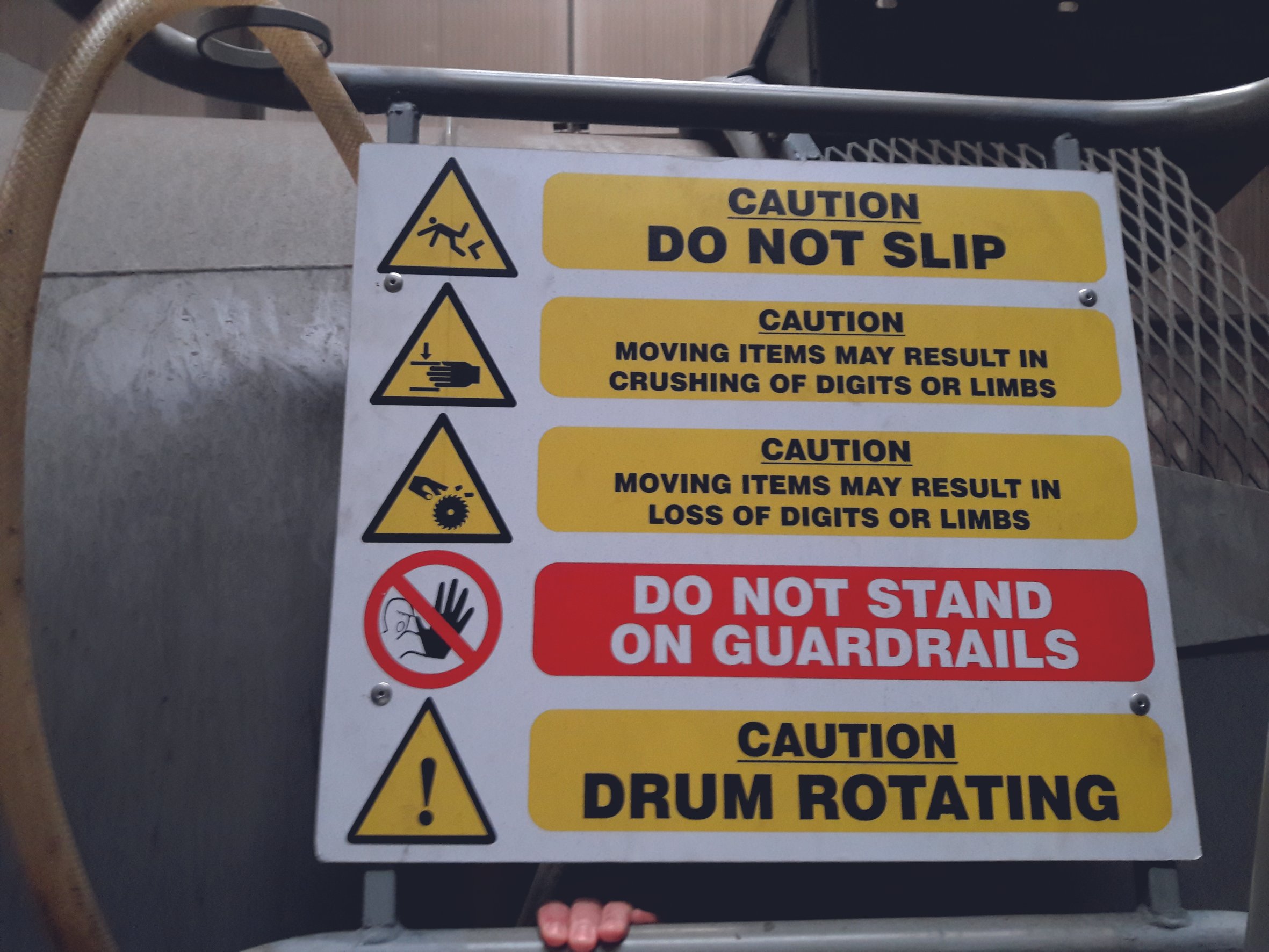A safety sign with various caution warnings including 'Do Not Slip,' 'Moving items may result in crushing of digits or limbs,' 'Moving items may result in loss of digits or limbs,' 'Do Not Stand on Guardrails,' and 'Drum Rotating.' The sign features