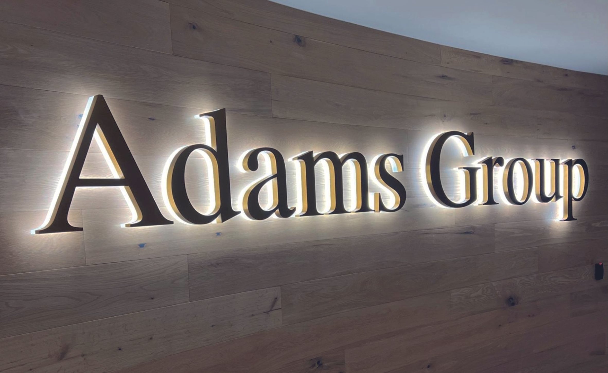 Illuminated "Adams Group" sign on a wood-paneled wall. Raised LED Letters. Illuminated signage. Signmakers Cork & Kerry, Ireland.