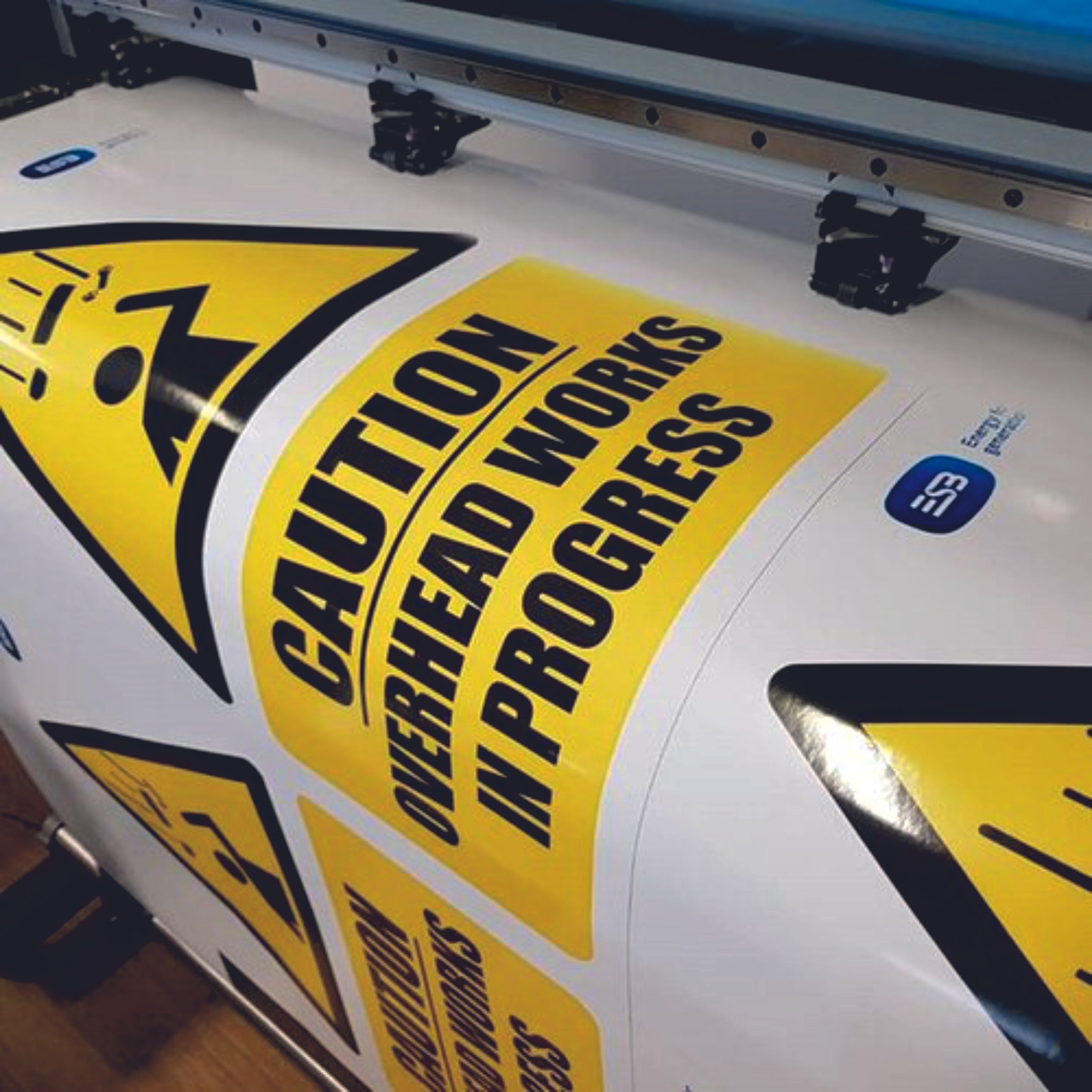 A printing machine producing caution signs with a triangular yellow background and the text 'CAUTION OVERHEAD WORKS IN PROGRESS.' The signs include a pictogram of a person and debris falling overhead, indicating a work hazard. Signmakers Cork & Kerry