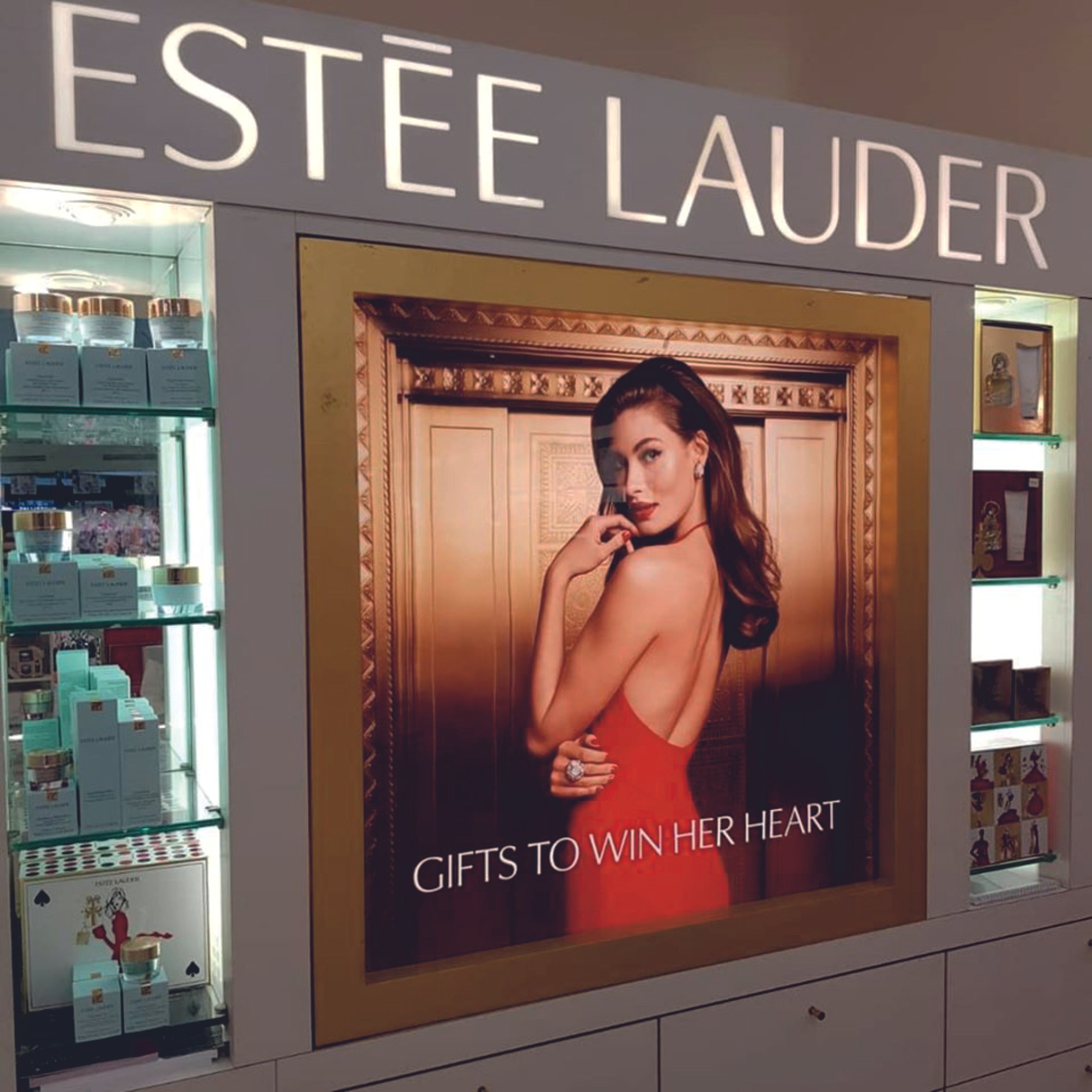 Estée Lauder storefront with a display, featuring a model in a red dress advertising beauty products, surrounded by shelves of skincare items. Raised LED Letters. Illuminated signage. Signmakers Cork & Kerry, Ireland.