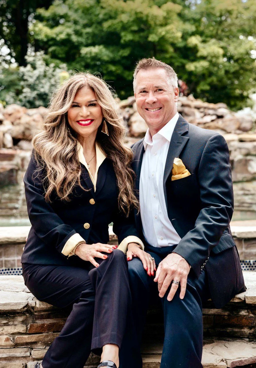 A smiling woman and man sitting together outdoors with greenery and rocks in the background, dressed in formal attire.