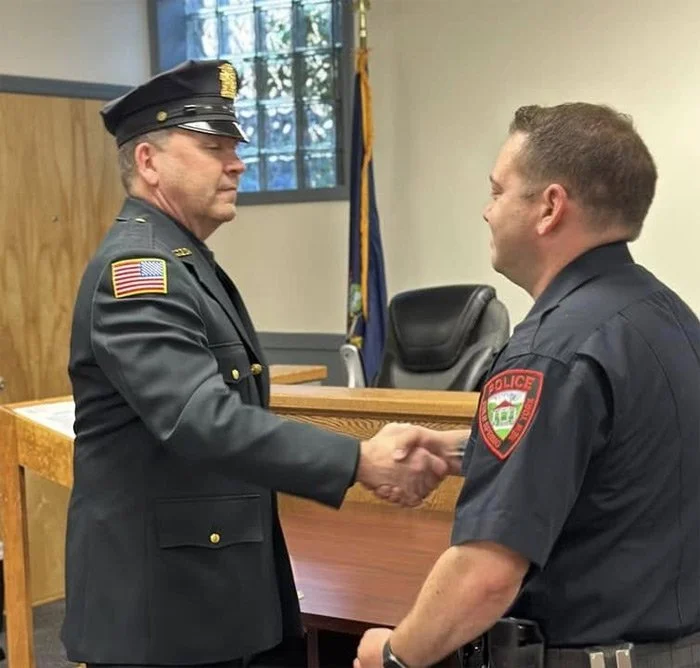 A uniformed officer with an American flag patch shakes hands with another officer in front of a judge's bench in a courtroom. The officer on the right has a police patch on his sleeve. They are smiling and exchanging greetings.