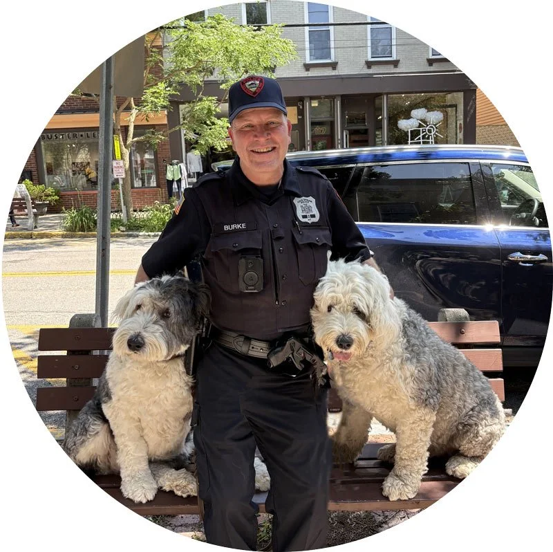 A police officer standing on a park bench with two large fluffy dogs, smiling. Behind them is a sunny street with storefronts and a black car parked nearby.