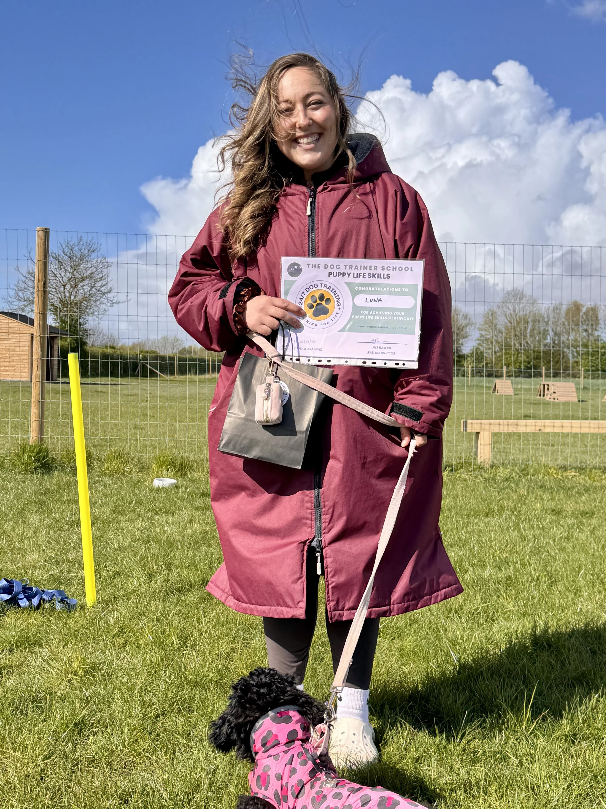 Young woman in a maroon coat holding a certificate and leash, sitting on grass with small black dog wearing a pink and black polka dot coat, outdoors with blue sky and white clouds. She has passed her puppy life skills course. 
