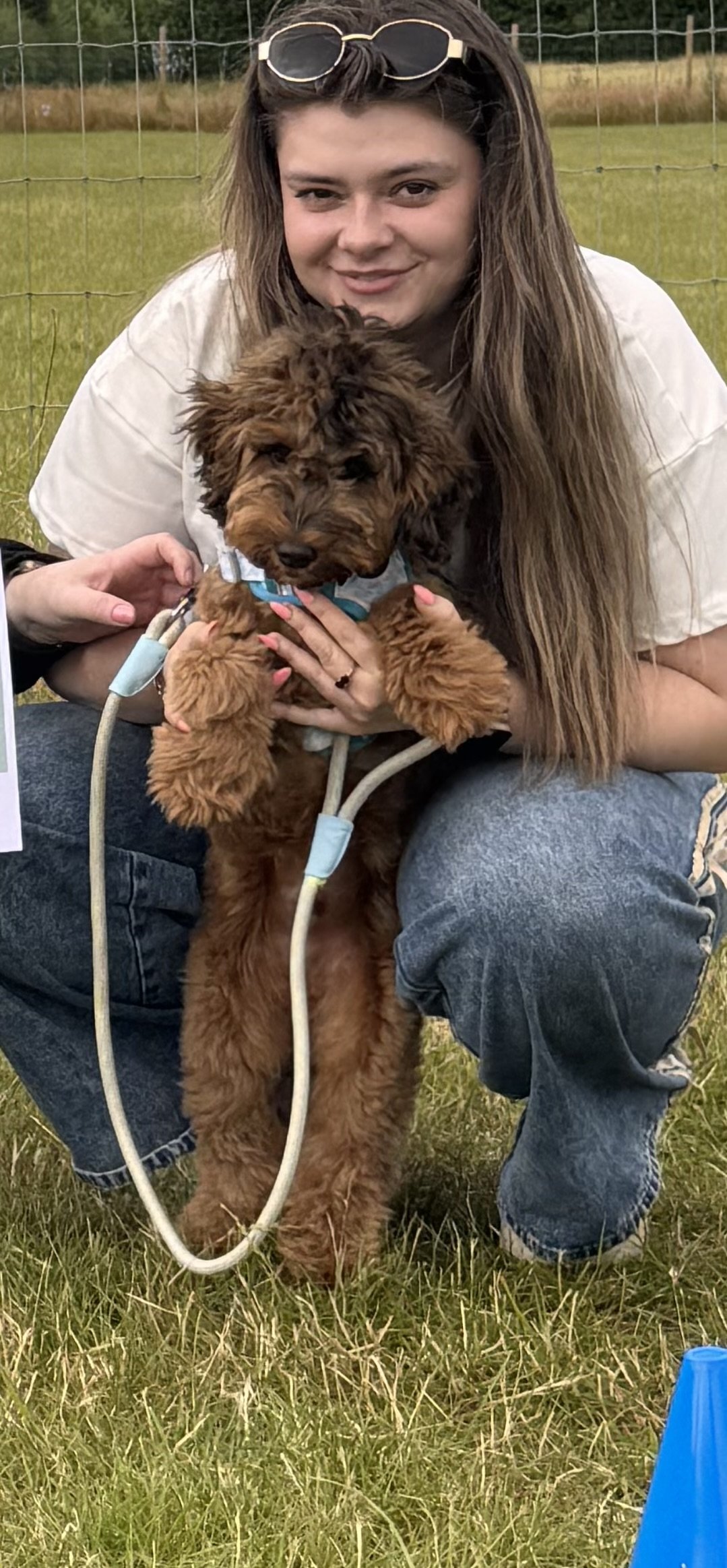 A woman with long brown hair, sunglasses on her head, smiling, holding a brown puppy with a red collar. They are outdoors in a grassy field with a fence in the background.