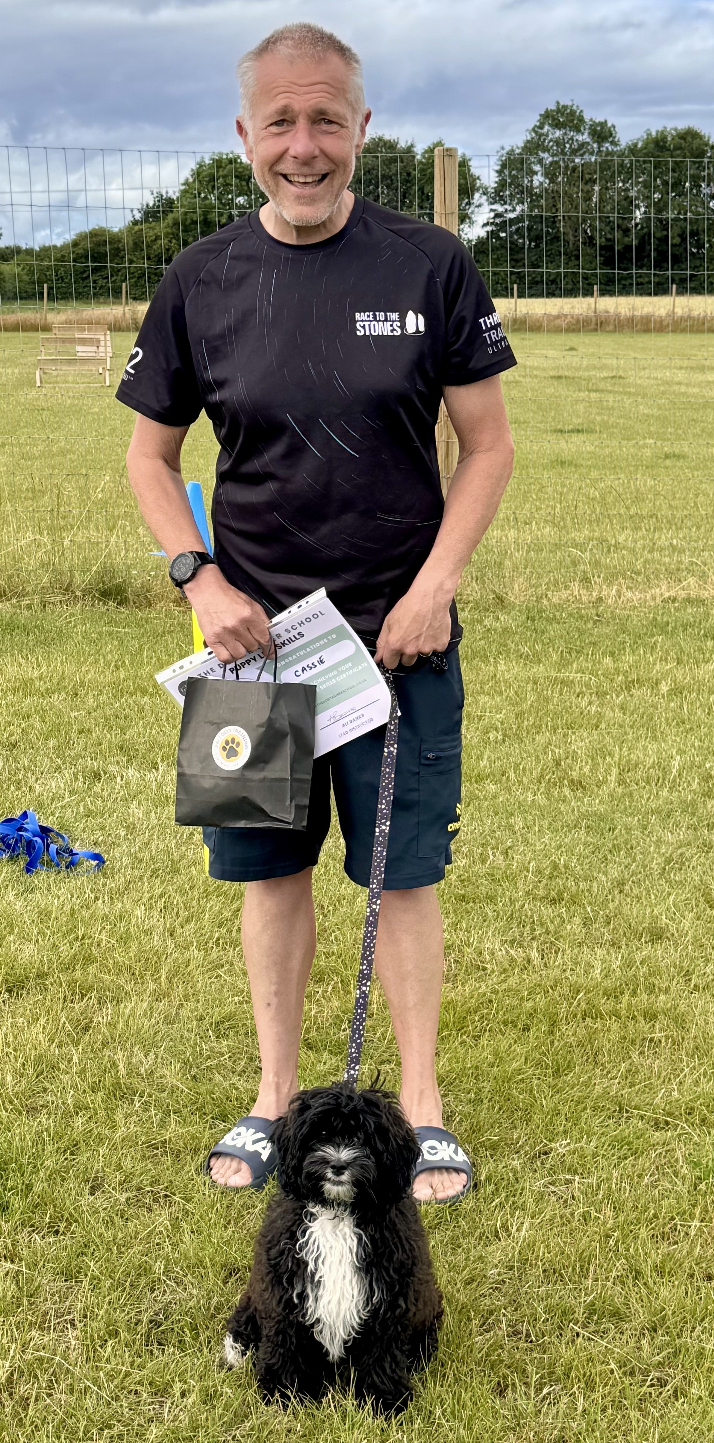 A man with short gray hair and a beard, smiling, stands on grass holding certificates and a black gift bag, with a small black and white dog in front of him. The man wears a black T-shirt with logos and black shorts, and the background features a wir