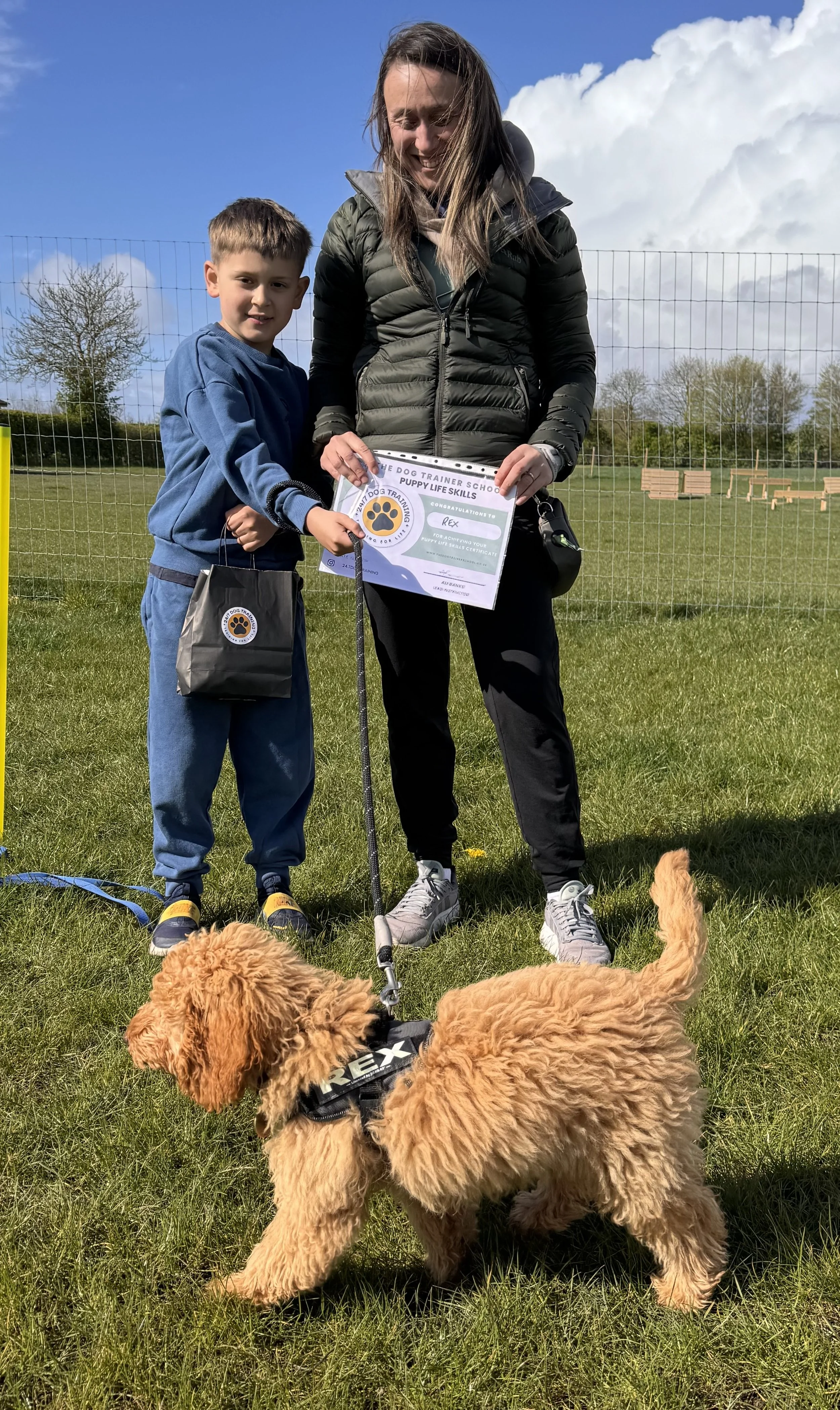 A woman and boy holding a certificate and bag, standing on grass with two fluffy golden retriever puppies in front of them during daytime. The background shows a fenced outdoor area with trees and benches under a partly cloudy sky.