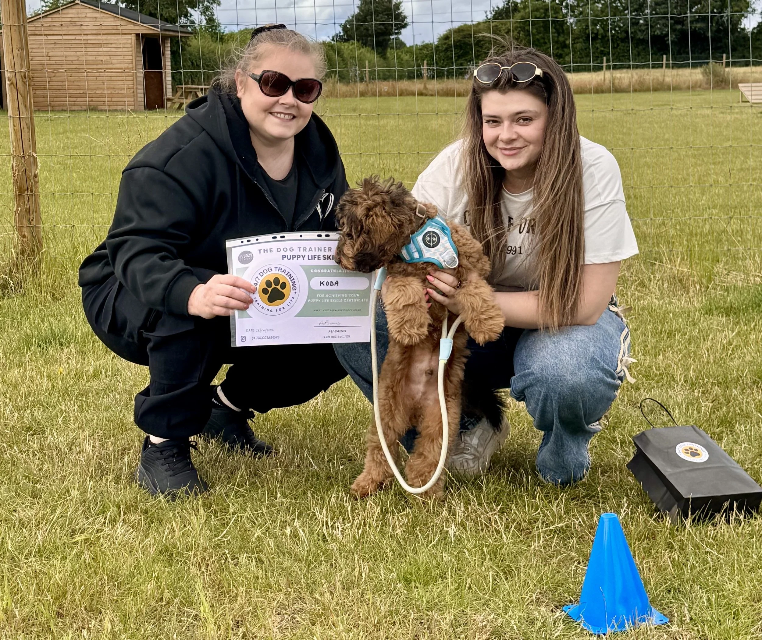 Two women kneeling on grass with a small brown puppy, holding a certificate and smiling, outdoors near a wooden structure and a field, with a blue cone marker nearby. In a private puppy training facility in Widnes