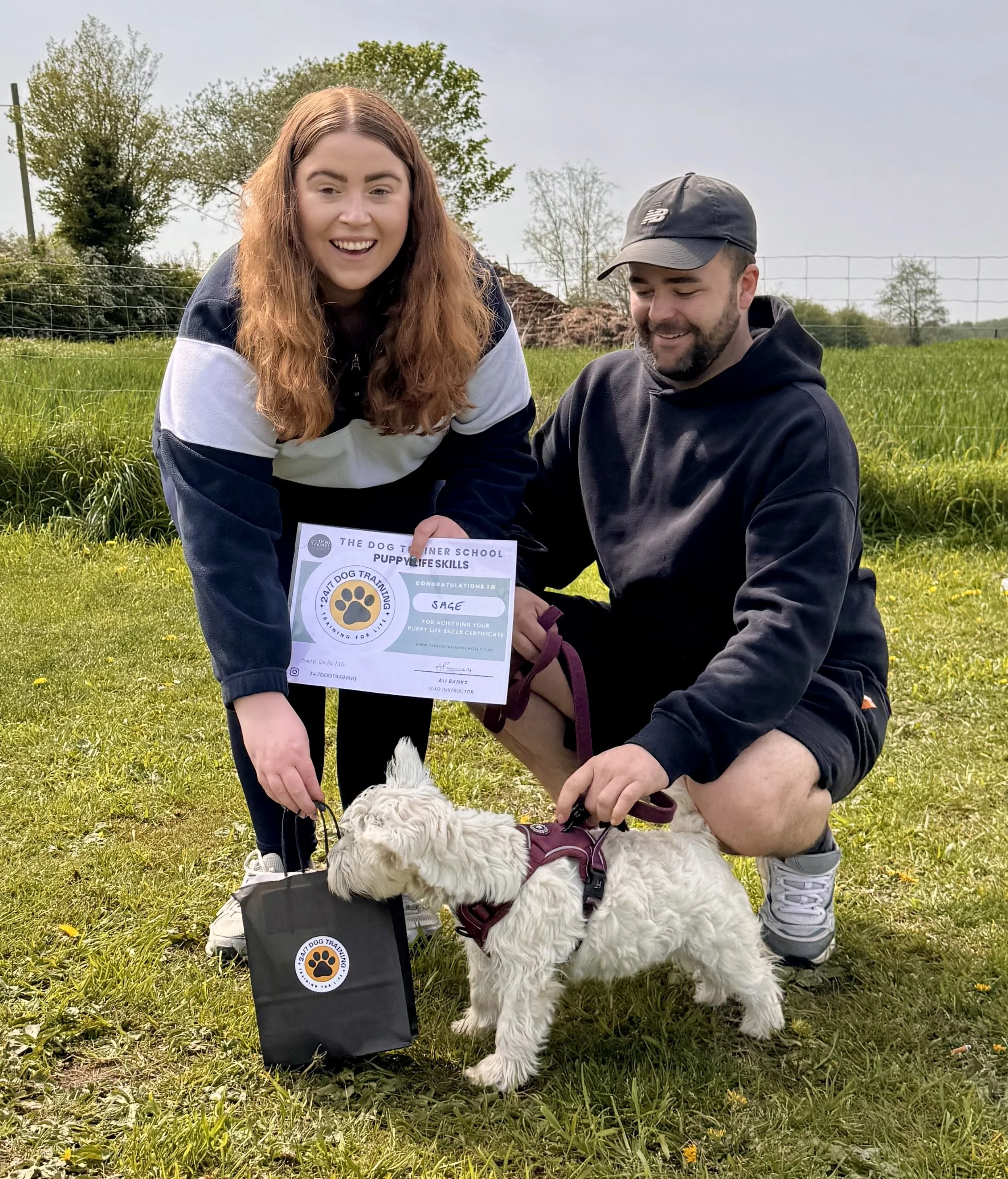 A woman with long red hair and a man with a beard and a cap are outdoors in a grassy field. The woman is holding a certificate, and the man is standing next to a fluffy white dog. They are smiling, celebrating a dog training achievement.