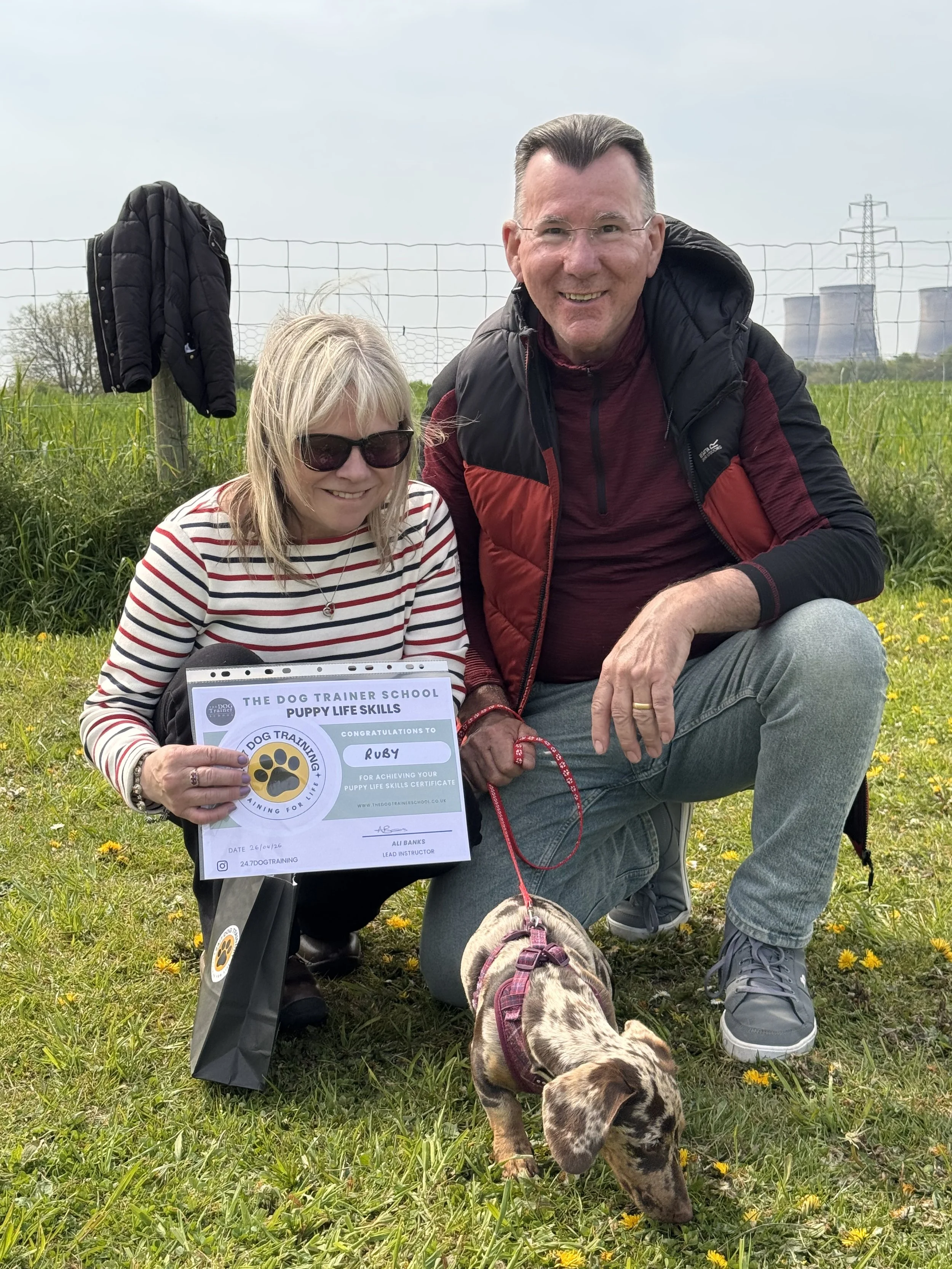 A woman with sunglasses holding a certificate from a dog training school, sitting on the grass with a brindle puppy on a leash, and a man beside her kneeling on the grass, in an outdoor field with yellow flowers and power plant cooling towers in the 