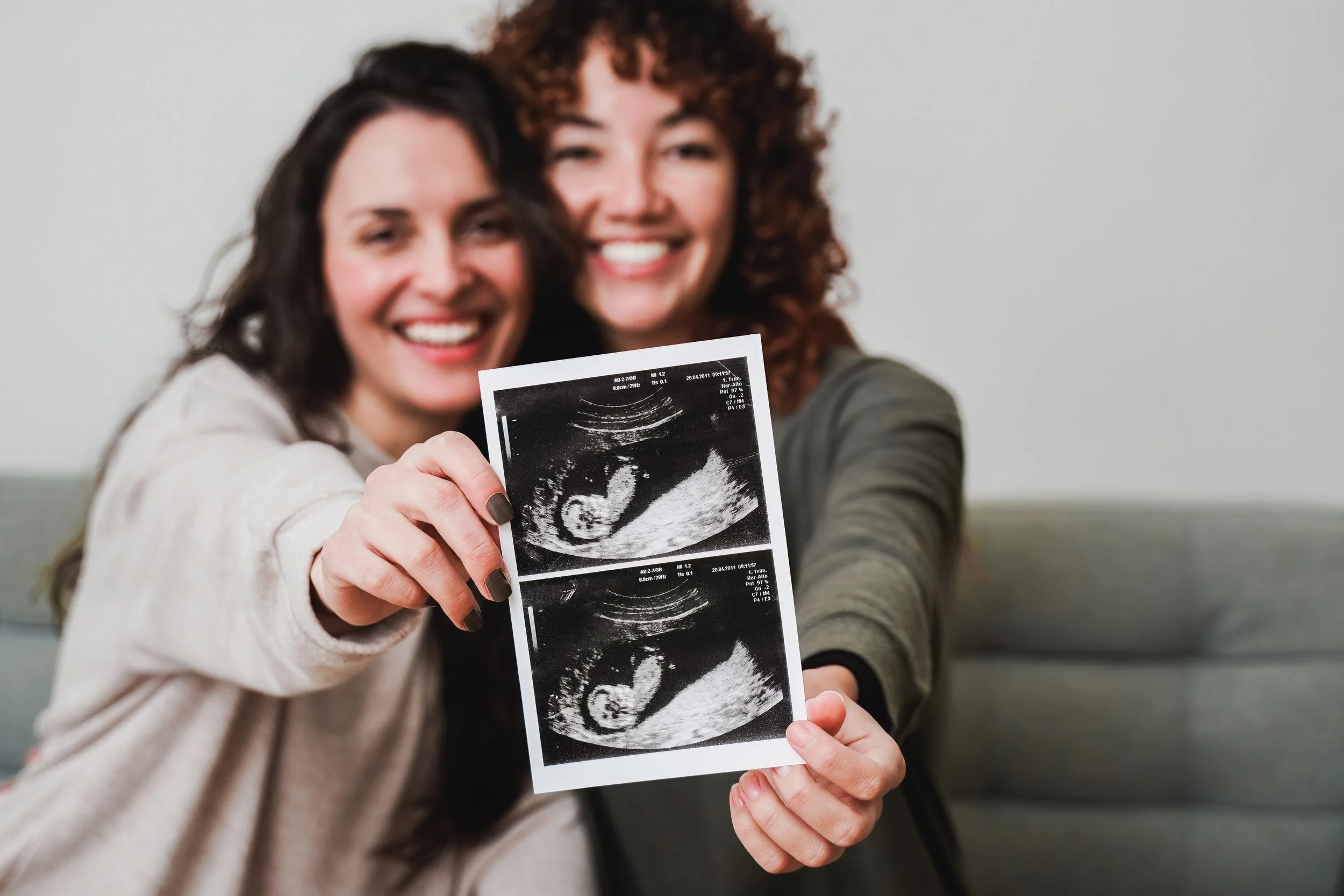 Two woman are side by side holding an ultrasound photo series together with big smiles on their faces.