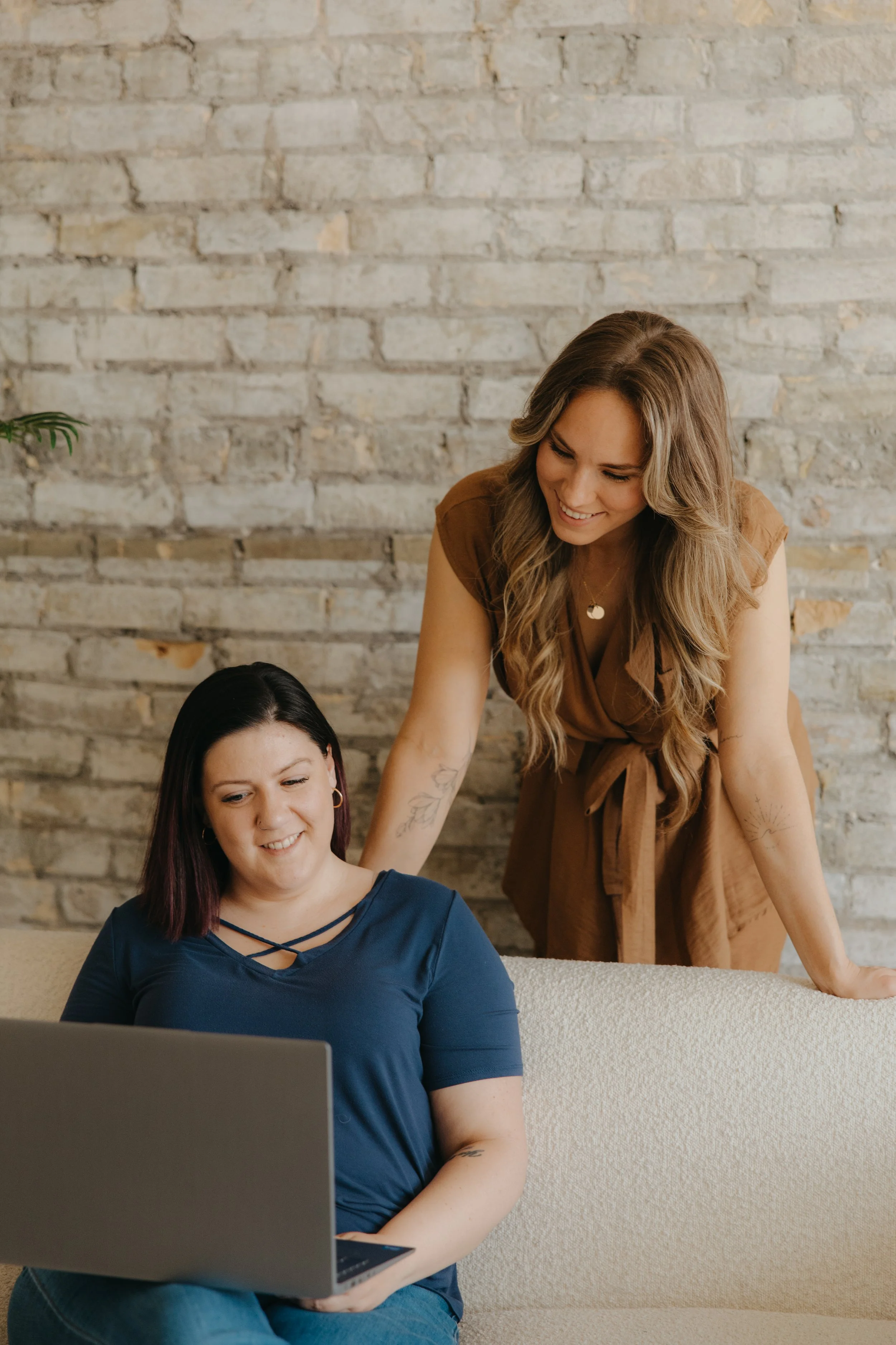 Two women smiling and looking at a laptop screen, one sitting on a couch and the other leaning over behind her, against a brick wall.