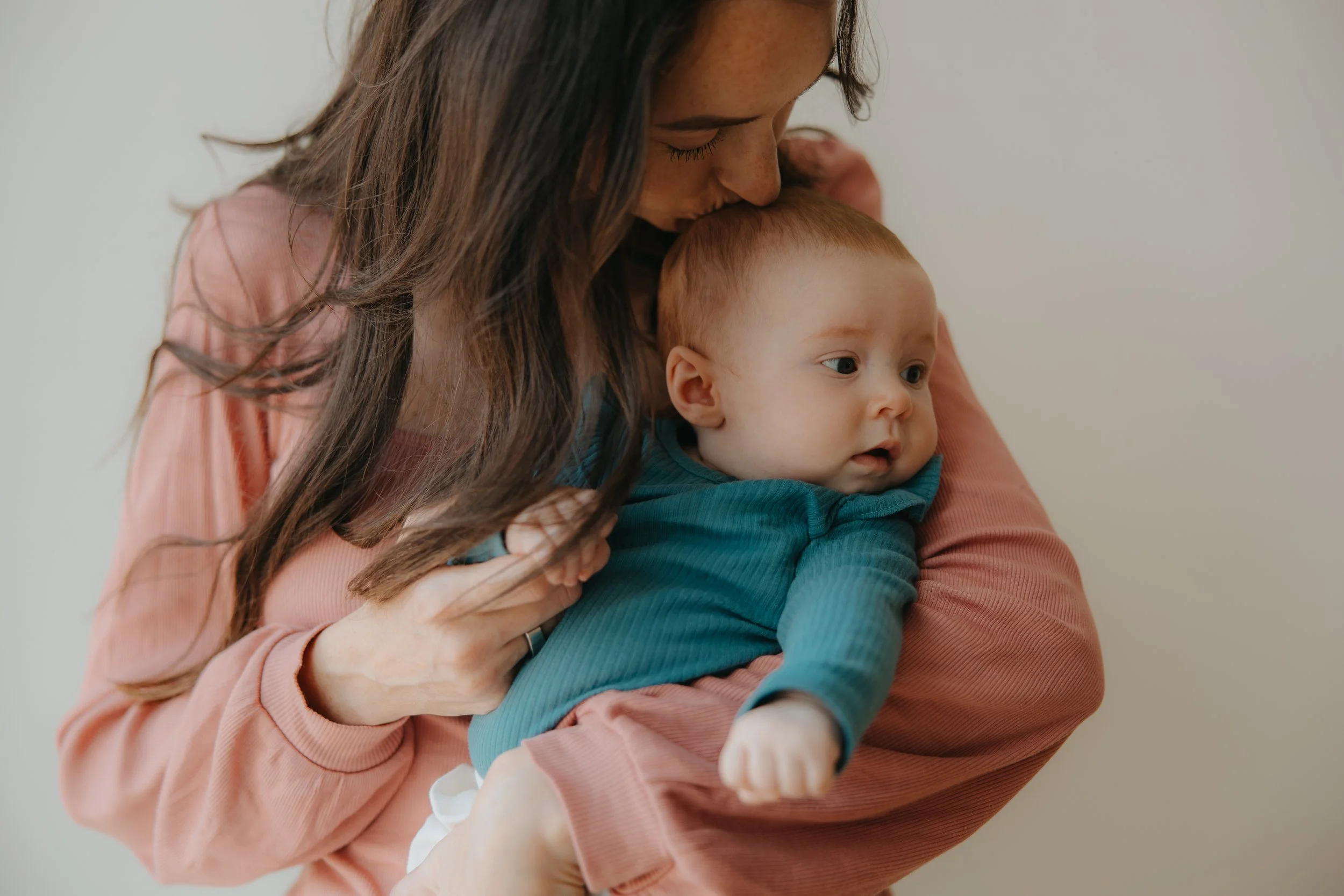 A woman with long dark hair holding a baby in a teal shirt and pink pants, as she kisses the baby's forehead.