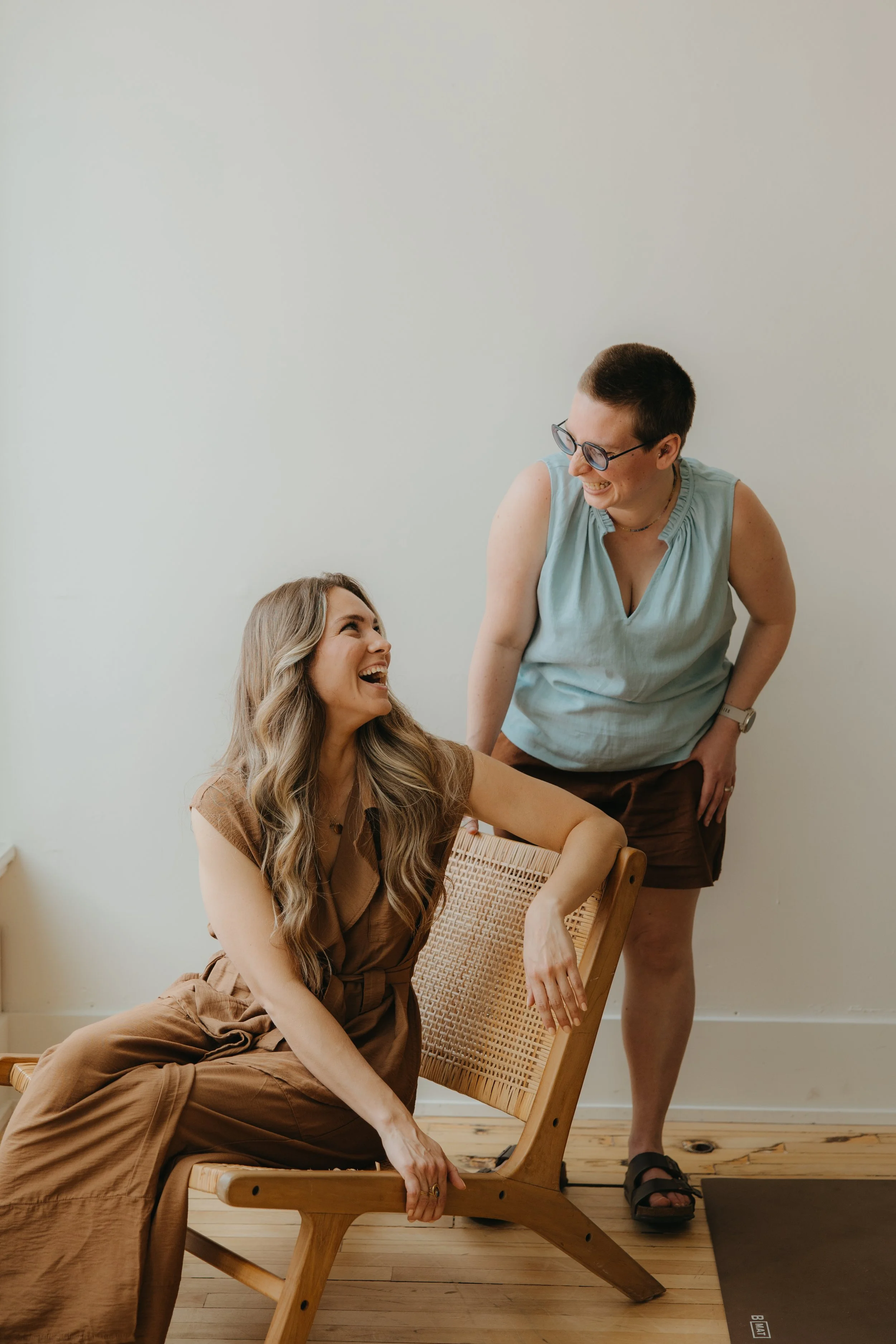 Two women sharing a laugh; one is seated in a wooden chair with a woven back, the other stands beside her.