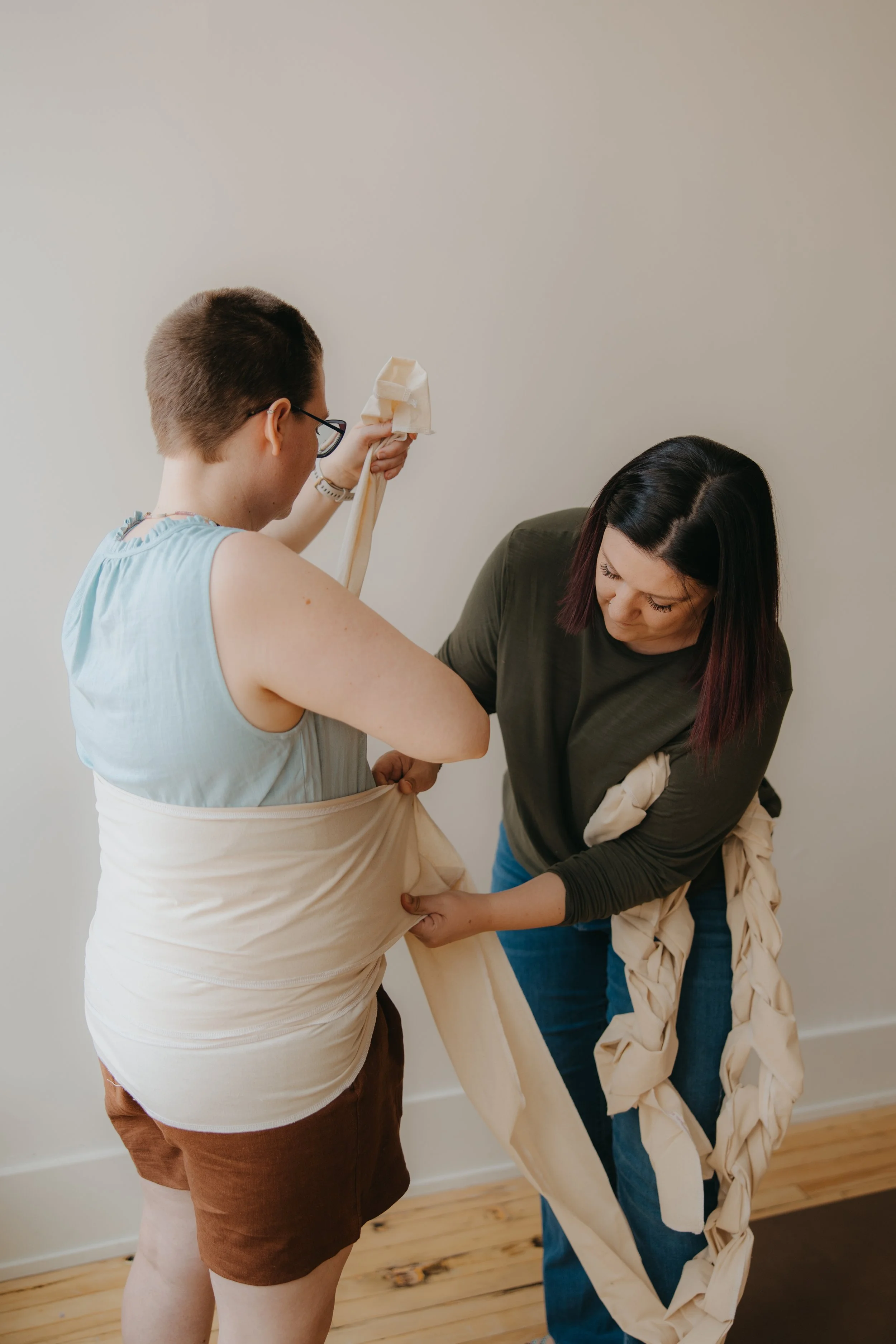 A doula with longer dark hair is wrapping a Benkung belly bind around the waist and abdomen of another woman with short brown hair and glasses. The woman having the bindings place on her is assisting by holding some of the beige fabric.
