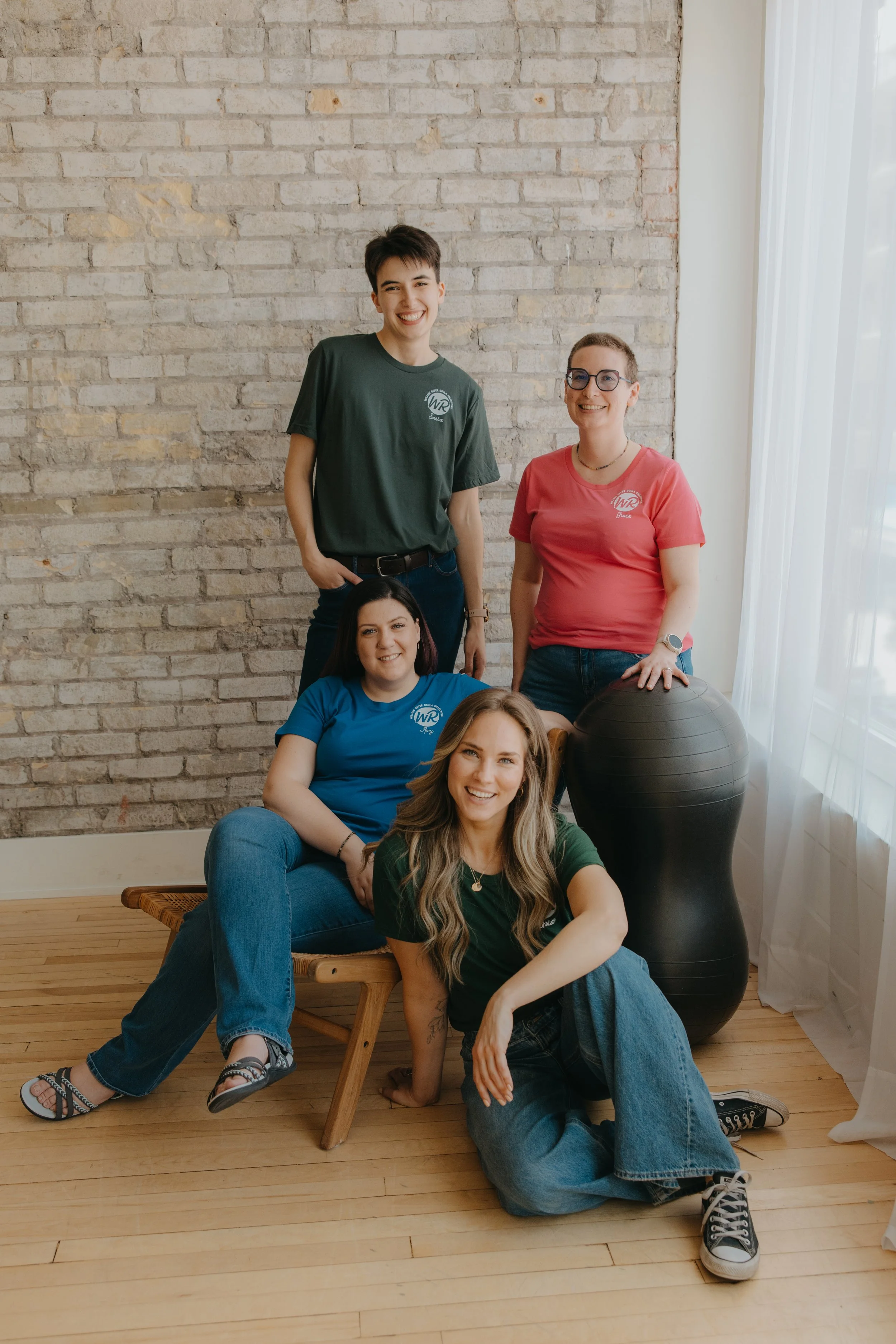 Group of four young women in colorful t-shirts, smiling, posed with a peanut ball indoors with a brick wall and sheer curtains in the background.