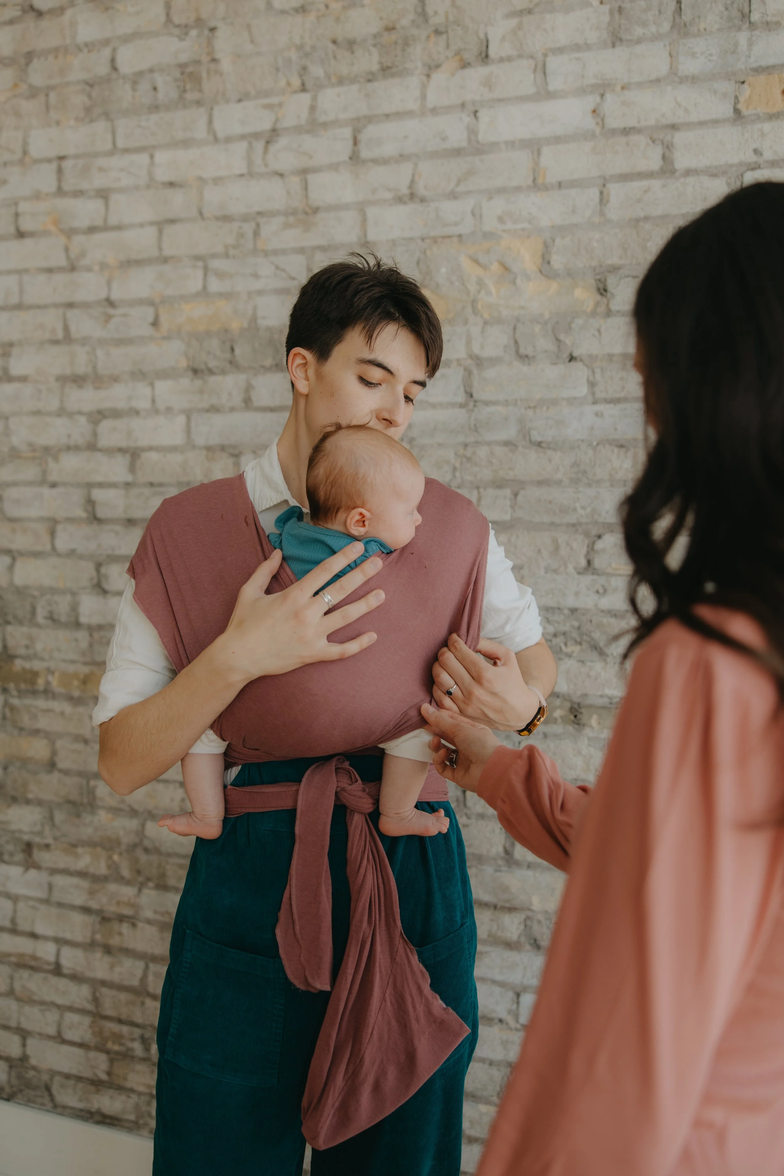 A doula with a baby in a baby carrier looking affectionately at the baby, next to a woman who is watching the demonstration.