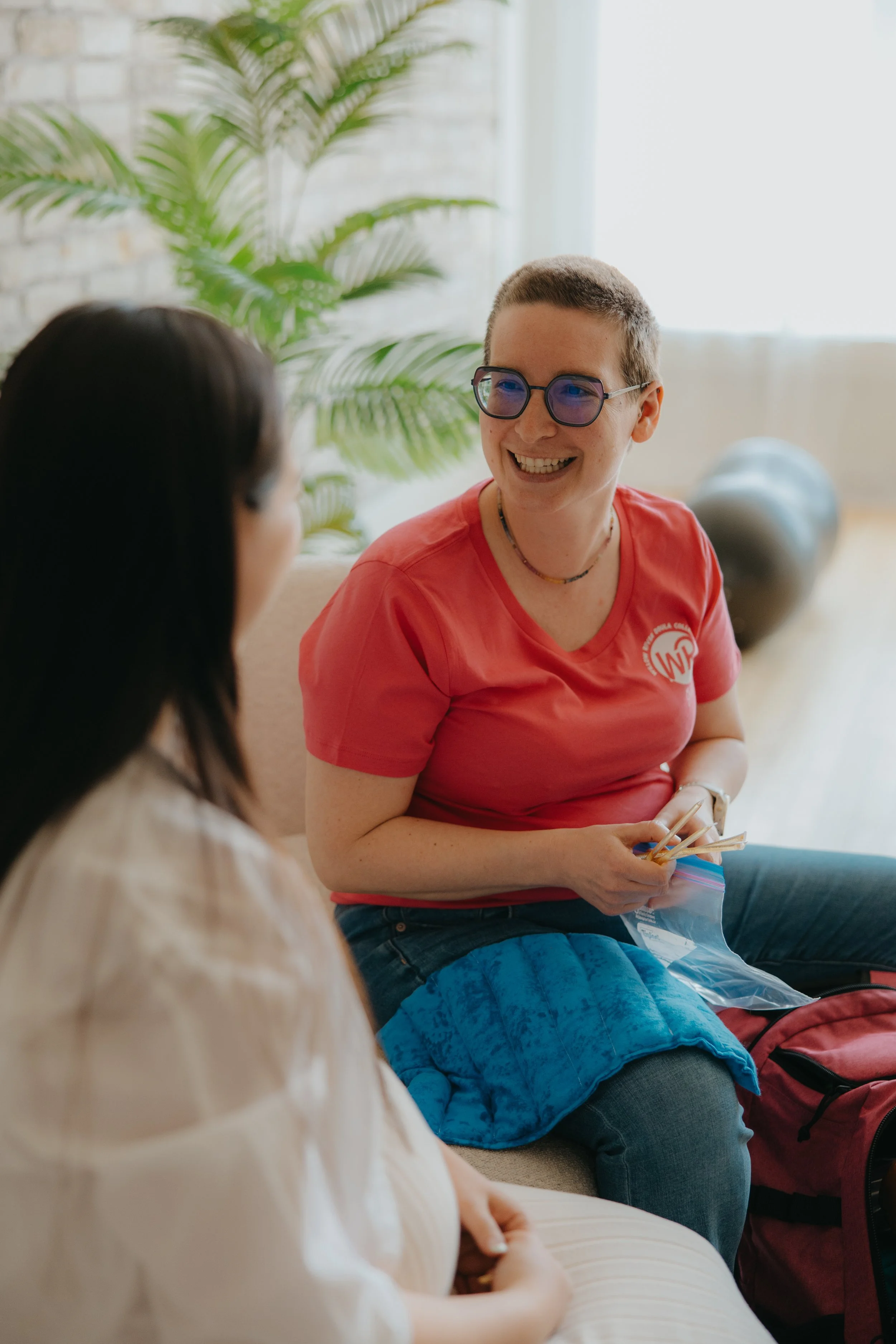 Two women having a conversation in a well-lit room with a large window, a houseplant, and a gray exercise ball in the background. One woman is wearing a red t-shirt and glasses, smiling, while the other has long dark hair and is wearing a beige top.