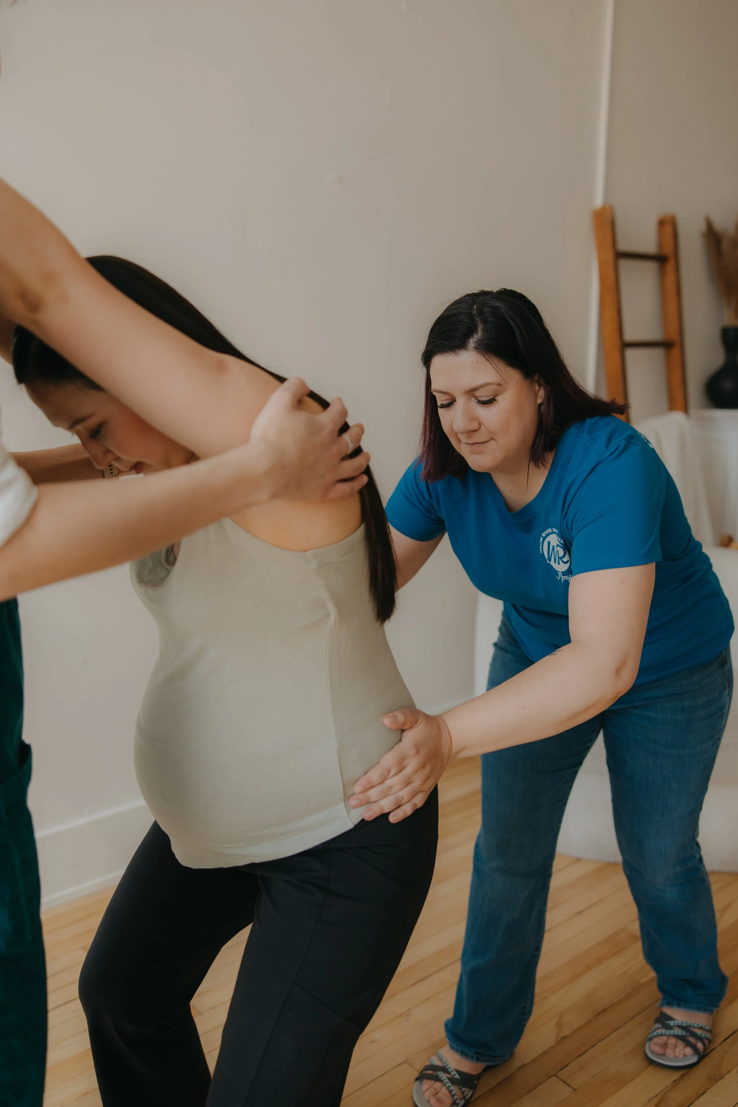 A woman in a blue shirt provides counter pressure to a pregnant woman's hips while the pregnant woman has her arms around another person who is holding her shoulders to support her standing.