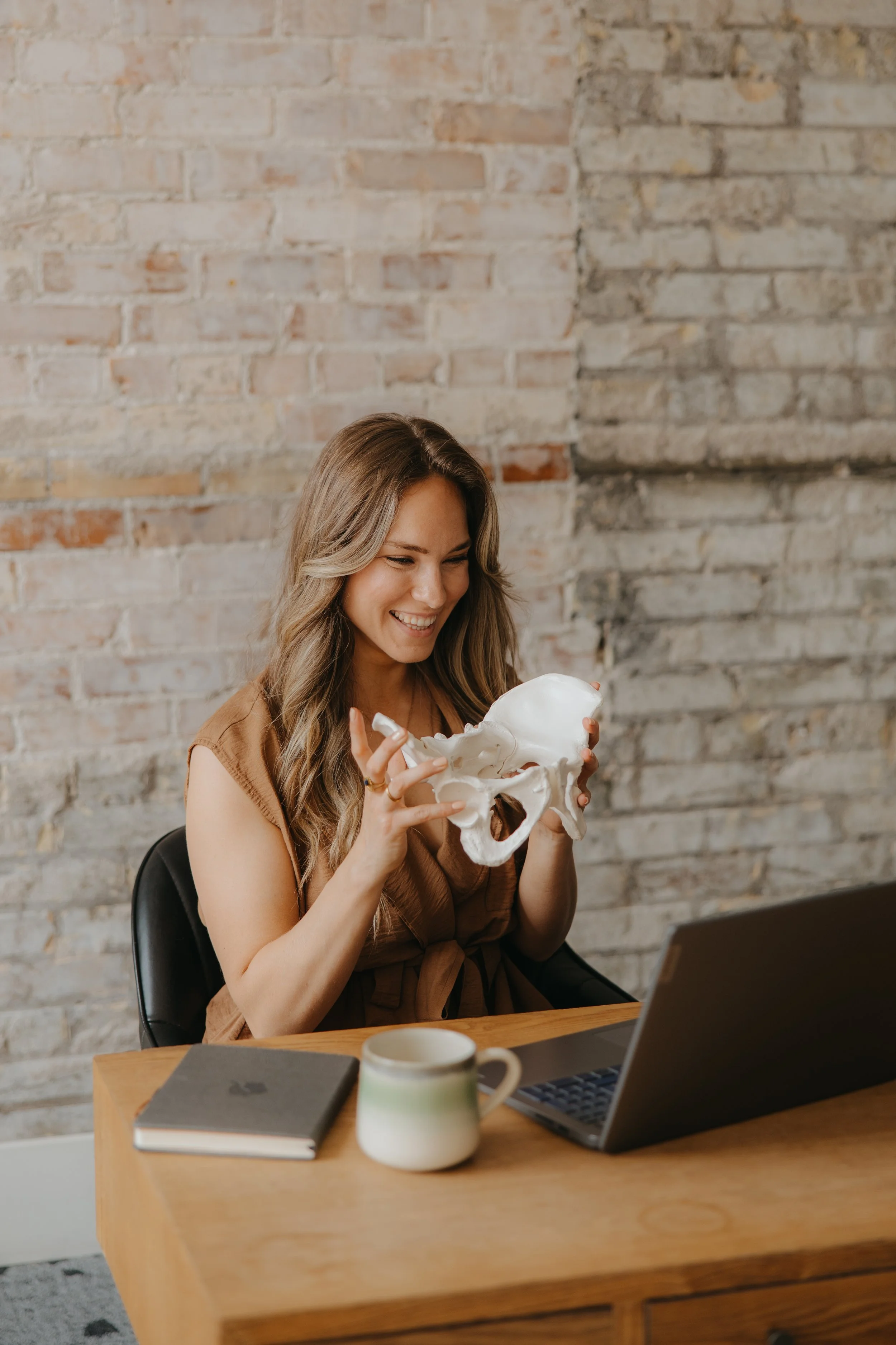A woman sitting at a wooden desk, holding a model of a human pelvis, smiling, with a laptop, a notebook, and a mug in front of her, against a brick wall background.