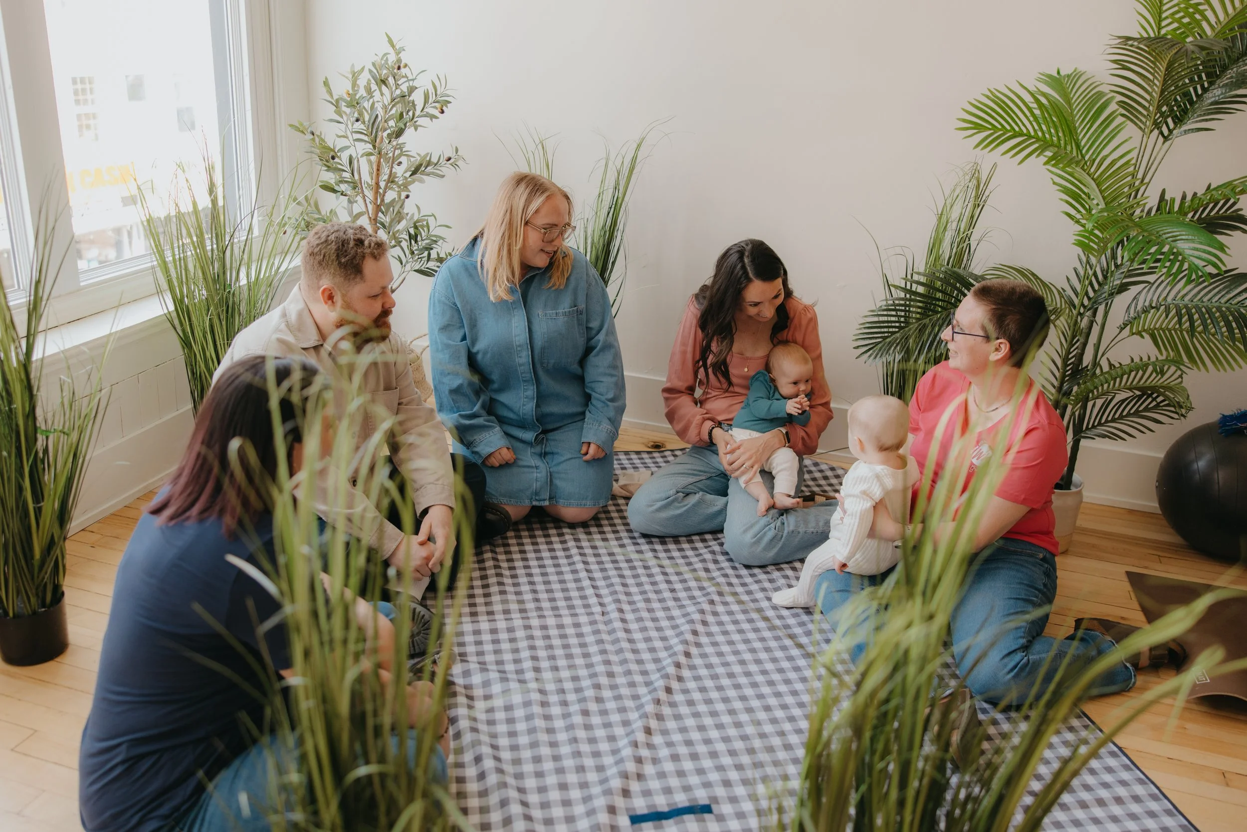 Group of adults and infants sitting on a checkered blanket inside a brightly lit room surrounded by tall green plants.