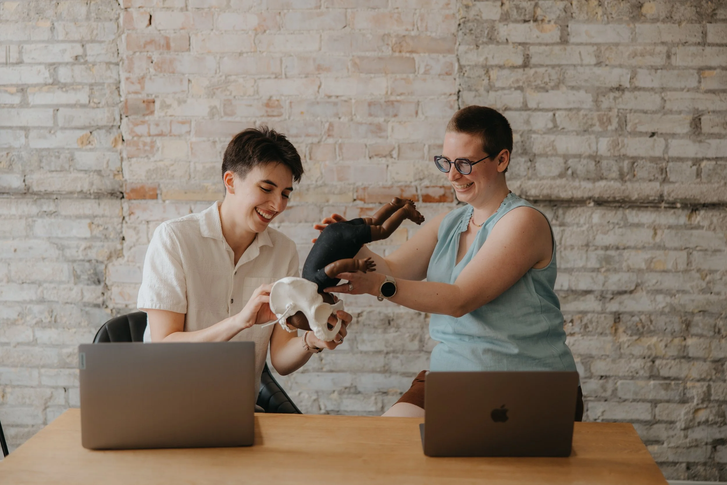Two women sitting at a table with laptops, smiling as they hold a model of a human pelvis and a small puppet of a baby demonstrating how the baby's head fits in the pelvis. The background is a white brick wall.