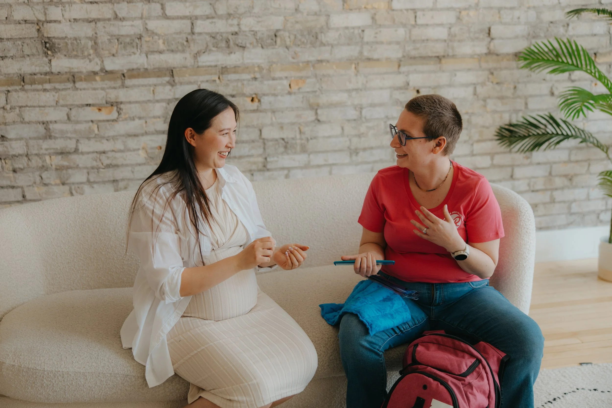 A pregnant woman sits on a couch with Grace who is wearing her pink WRDC tshirt and has her doula bag open with comfort tools in hand. They look engaged in conversation.