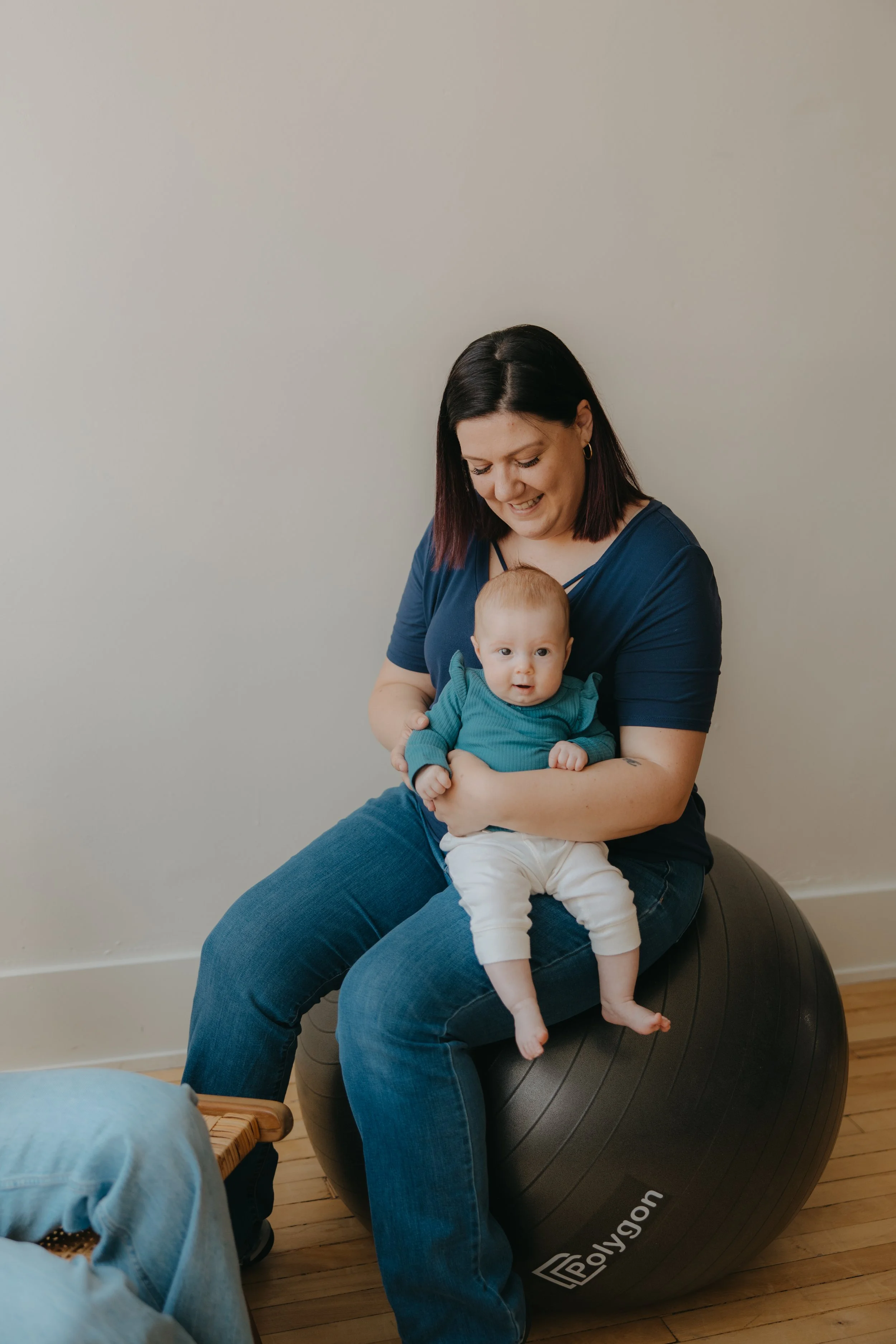 A woman with dark hair, wearing a blue shirt and jeans, sitting on a black exercise ball in a room with wooden floors, holding a young baby dressed in a teal sweater and white pants. The woman is smiling and looking down at the baby, who is looking a