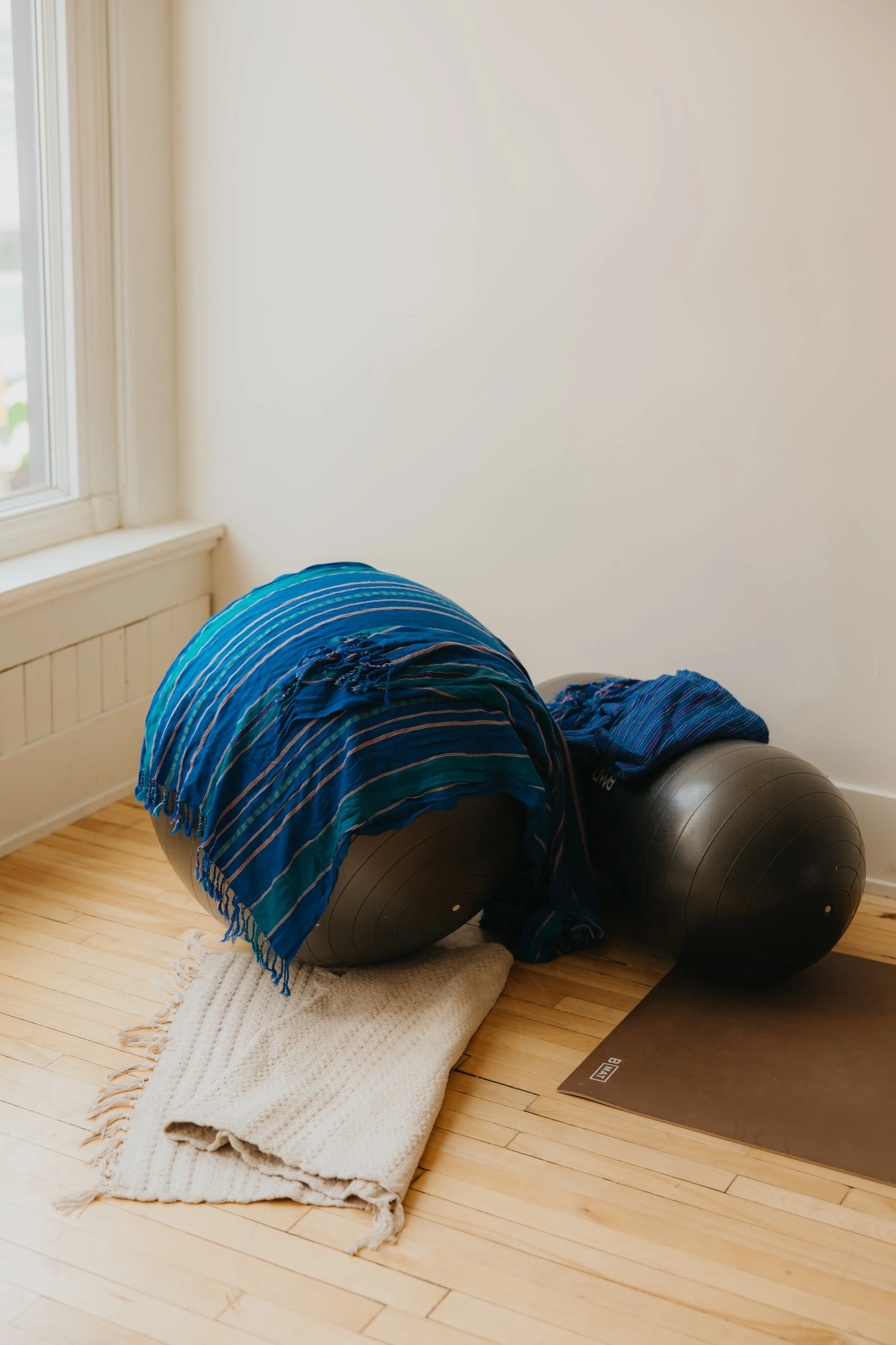 One birth ball and one peanut ball posed with a blue and green striped Rebozo draped over one, resting on a white rug in front of a window on a hardwood floor, next to a black yoga mat.